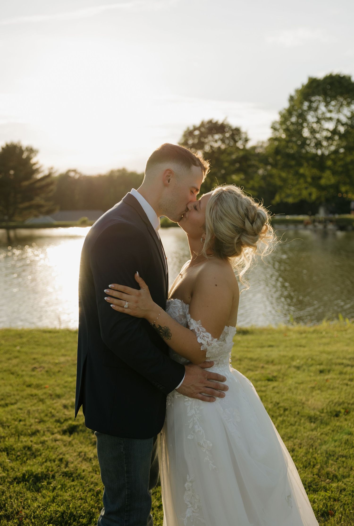 A bride and groom share a romantic kiss at golden hour by a serene pond, surrounded by lush greenery.