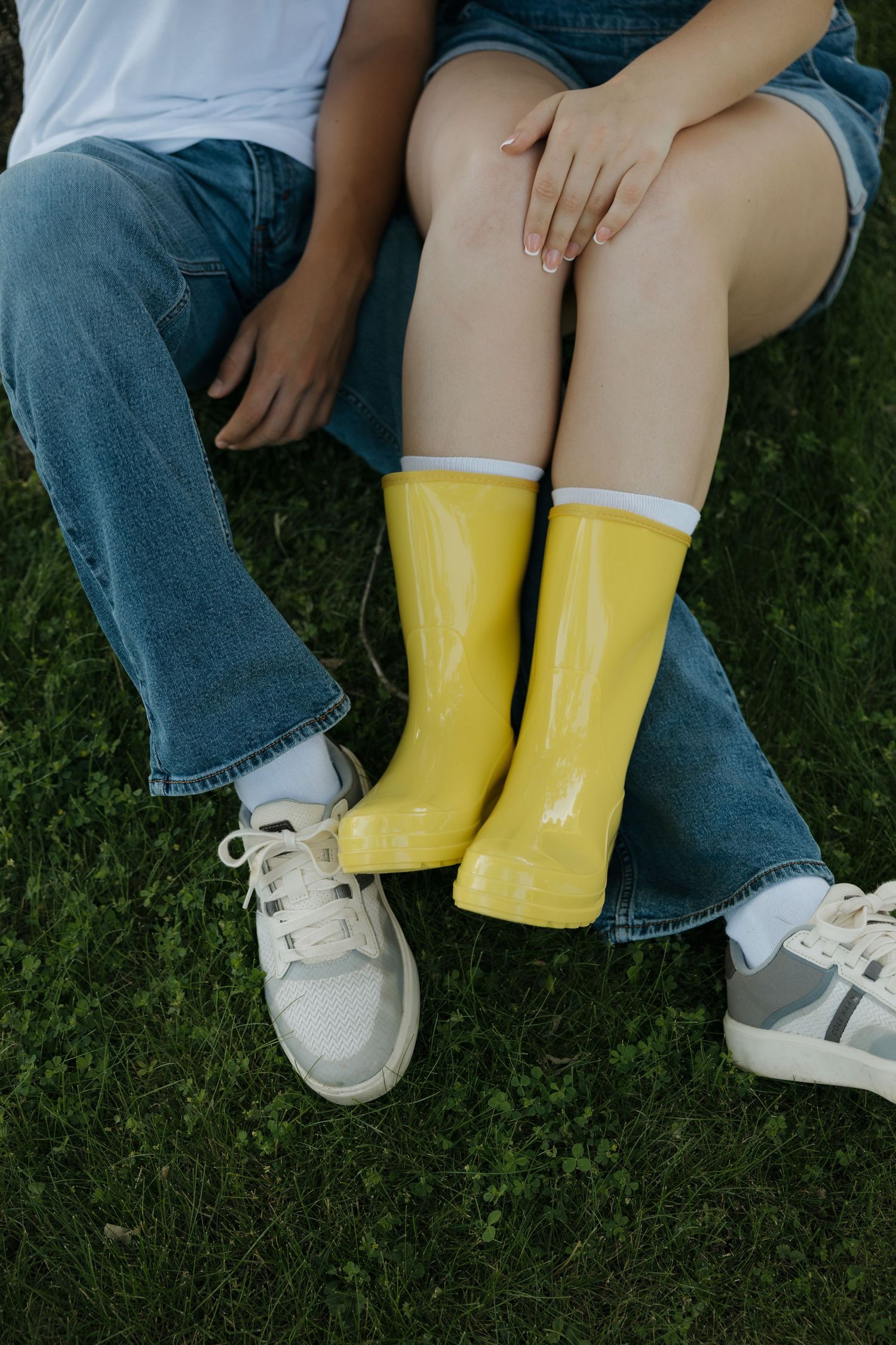 Two children sitting on grass, one wearing bright yellow rain boots and denim shorts, the other in sneakers and jeans.