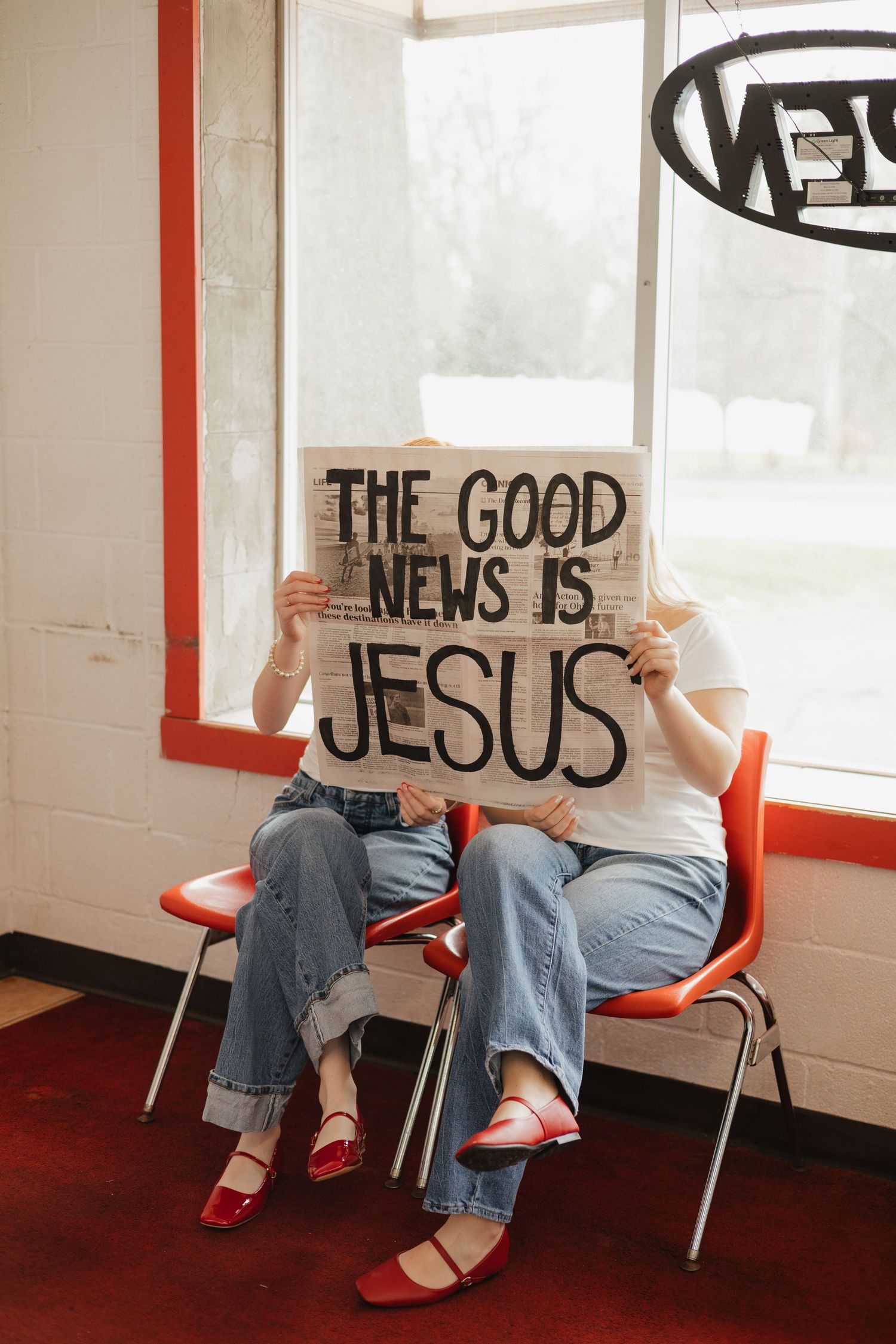 Two people in jeans sit on red chairs holding a cardboard sign that reads 'THE GOOD NEWS IS JESUS' in a bright room.