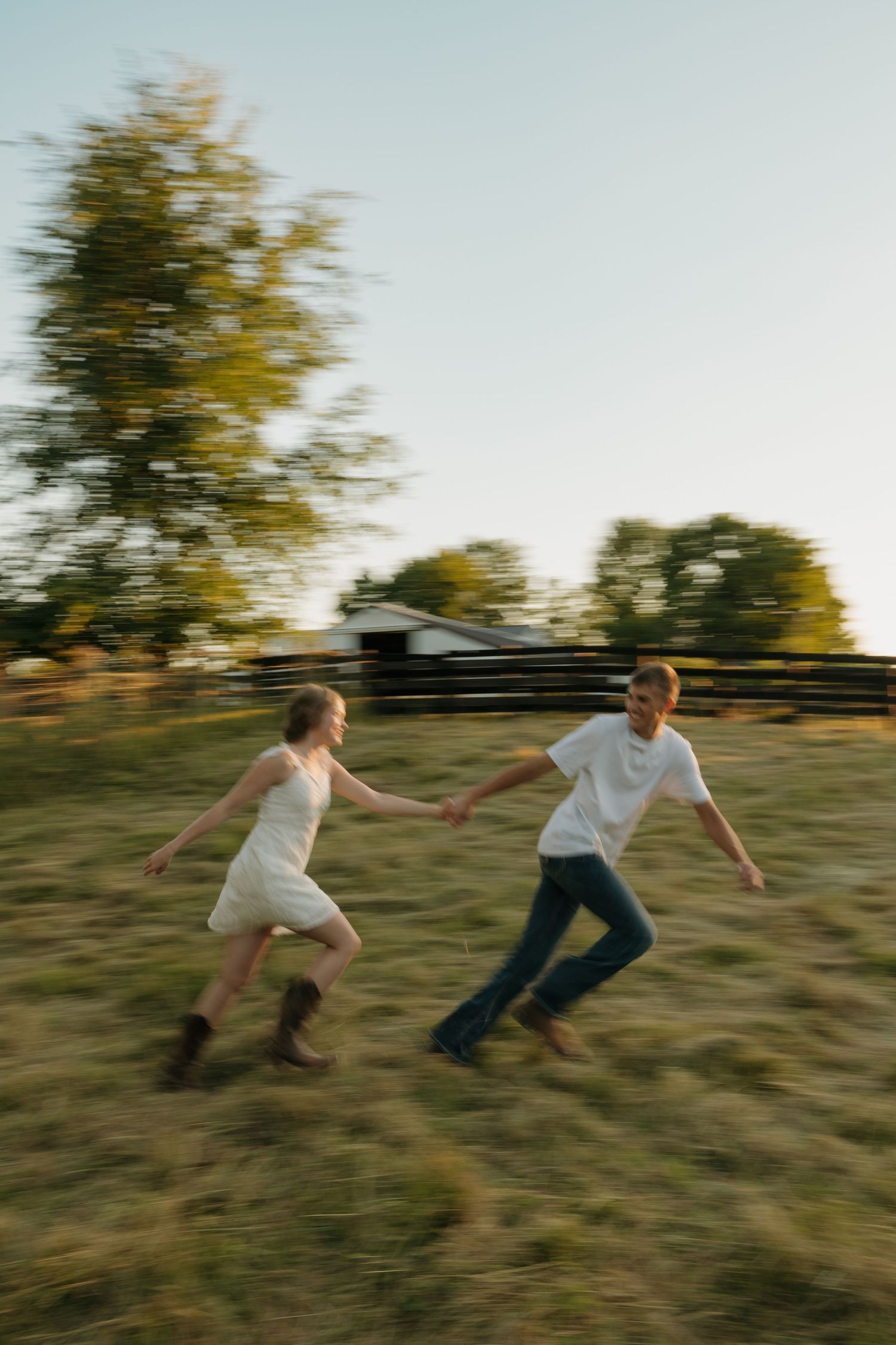 Joyful couple holding hands running through a green field at golden hour near a rustic farm fence.