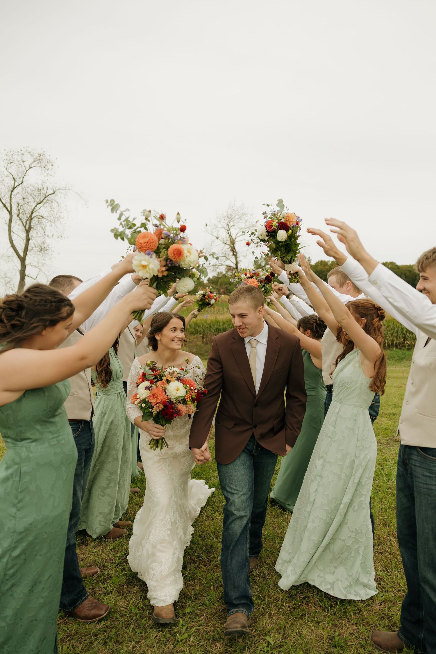 Bride and groom walk hand in hand through an arch of bouquets held by bridesmaids in sage green dresses at an outdoor wedding.