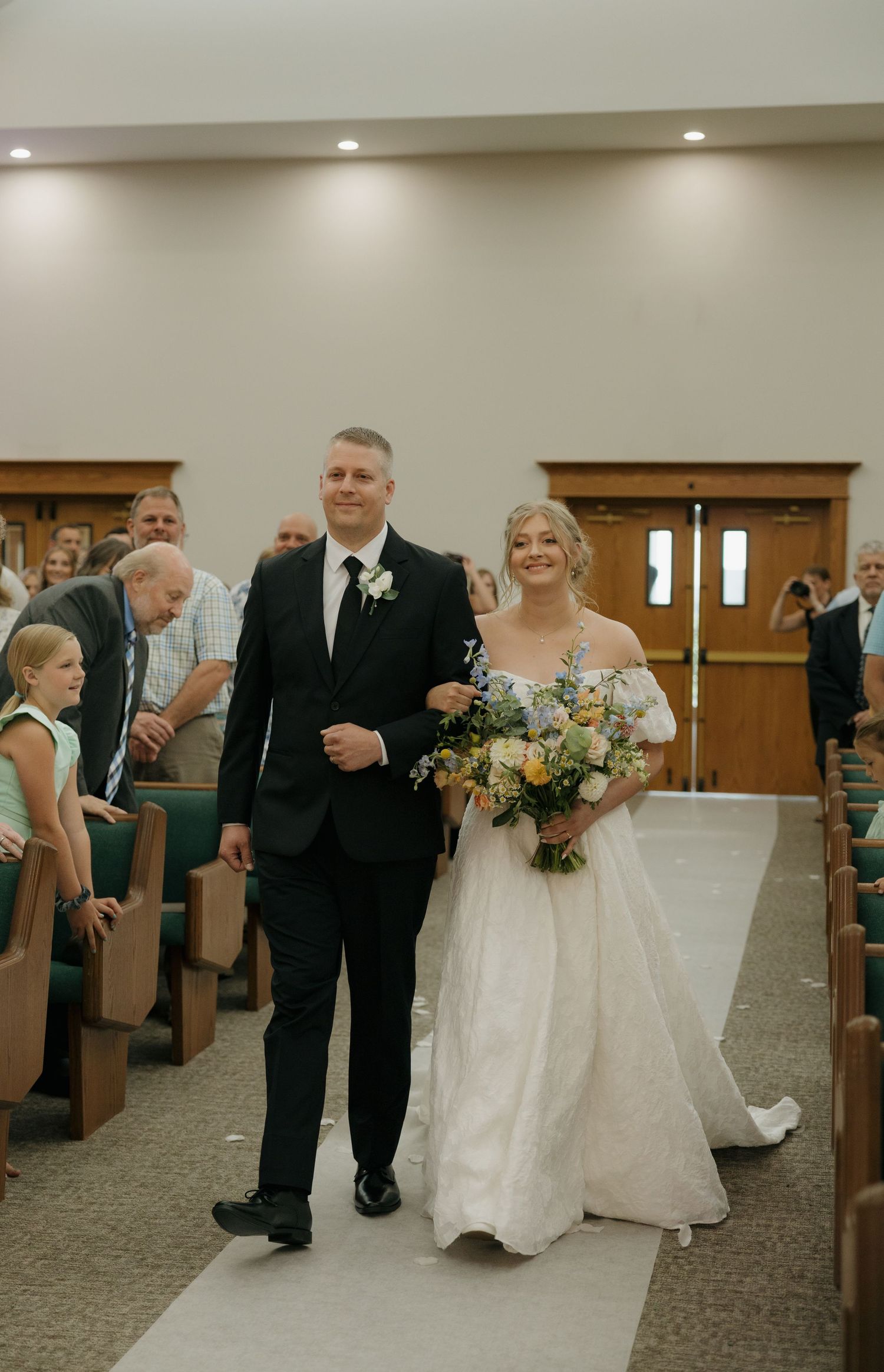Bride and groom walk down church aisle after wedding ceremony, bride holding colorful bouquet in elegant white gown.