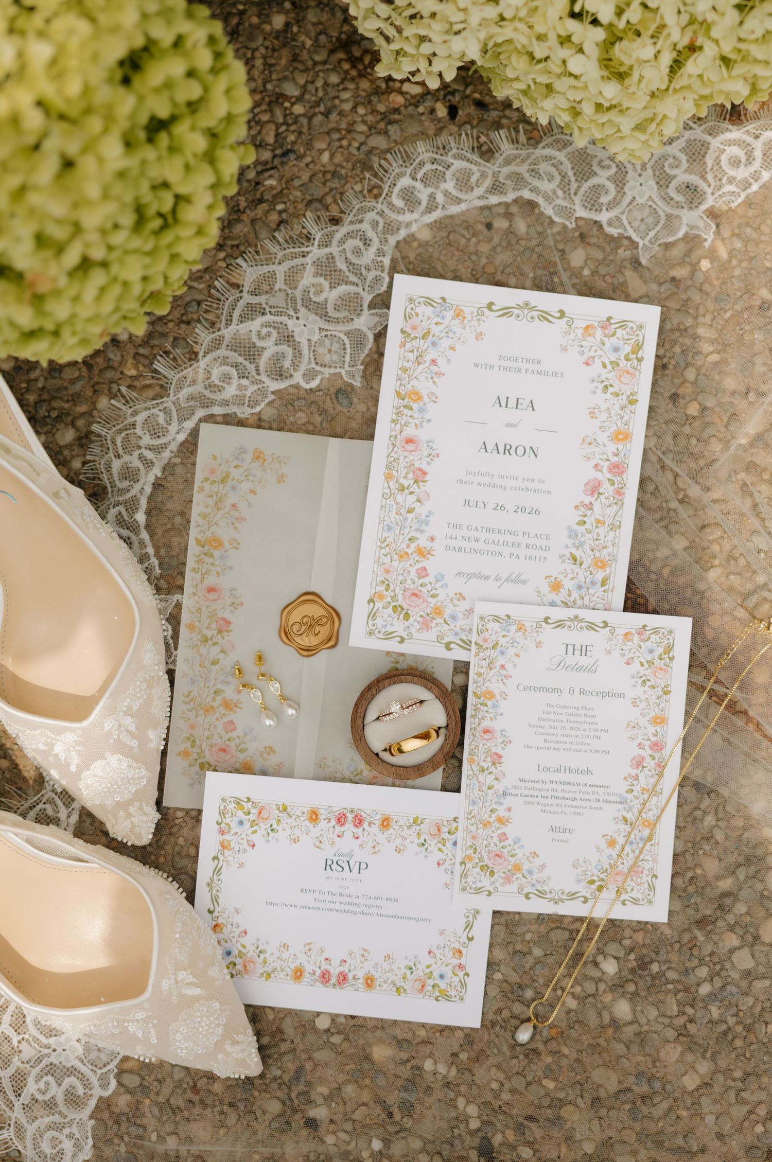 Elegant floral wedding invitation suite with RSVP cards, gold wax seal, rings, bridal heels, and lace veil on stone surface.