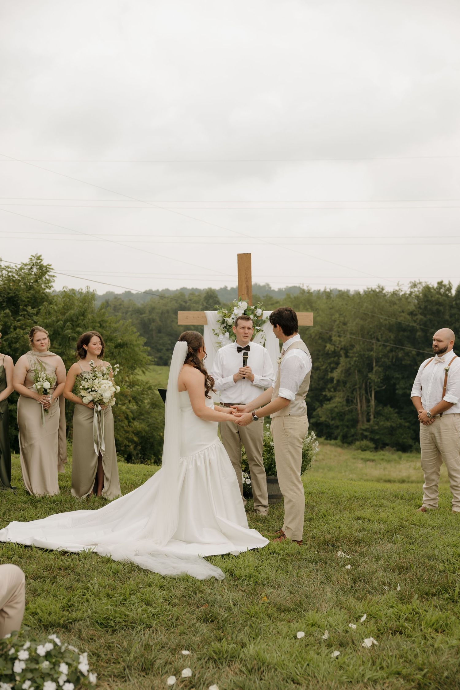 Outdoor wedding ceremony on a green hillside with bride and groom exchanging vows before a wooden cross altar.