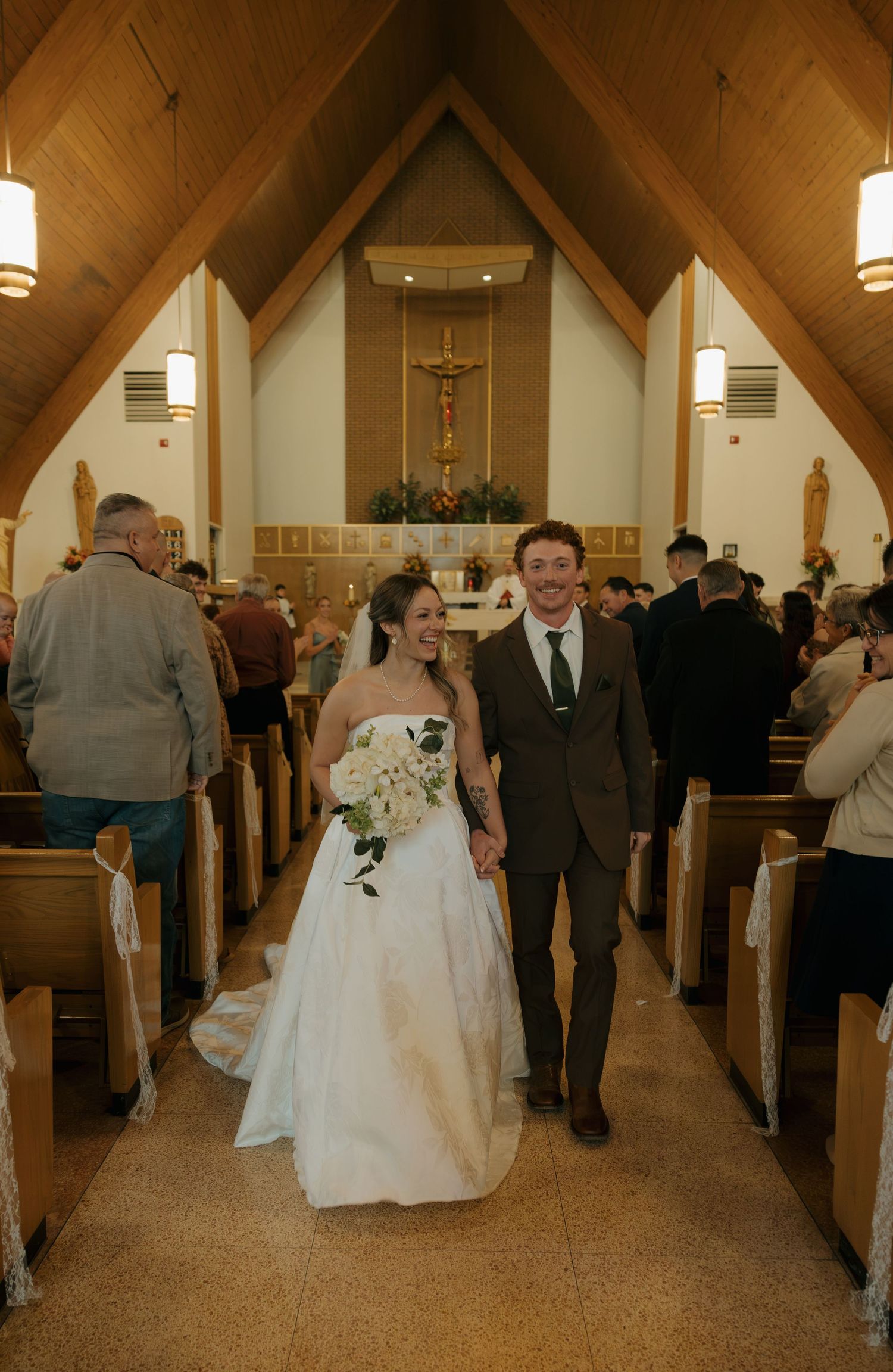 Smiling bride in a white gown holding a bouquet walks hand in hand with the groom down the church aisle past seated wedding guests.