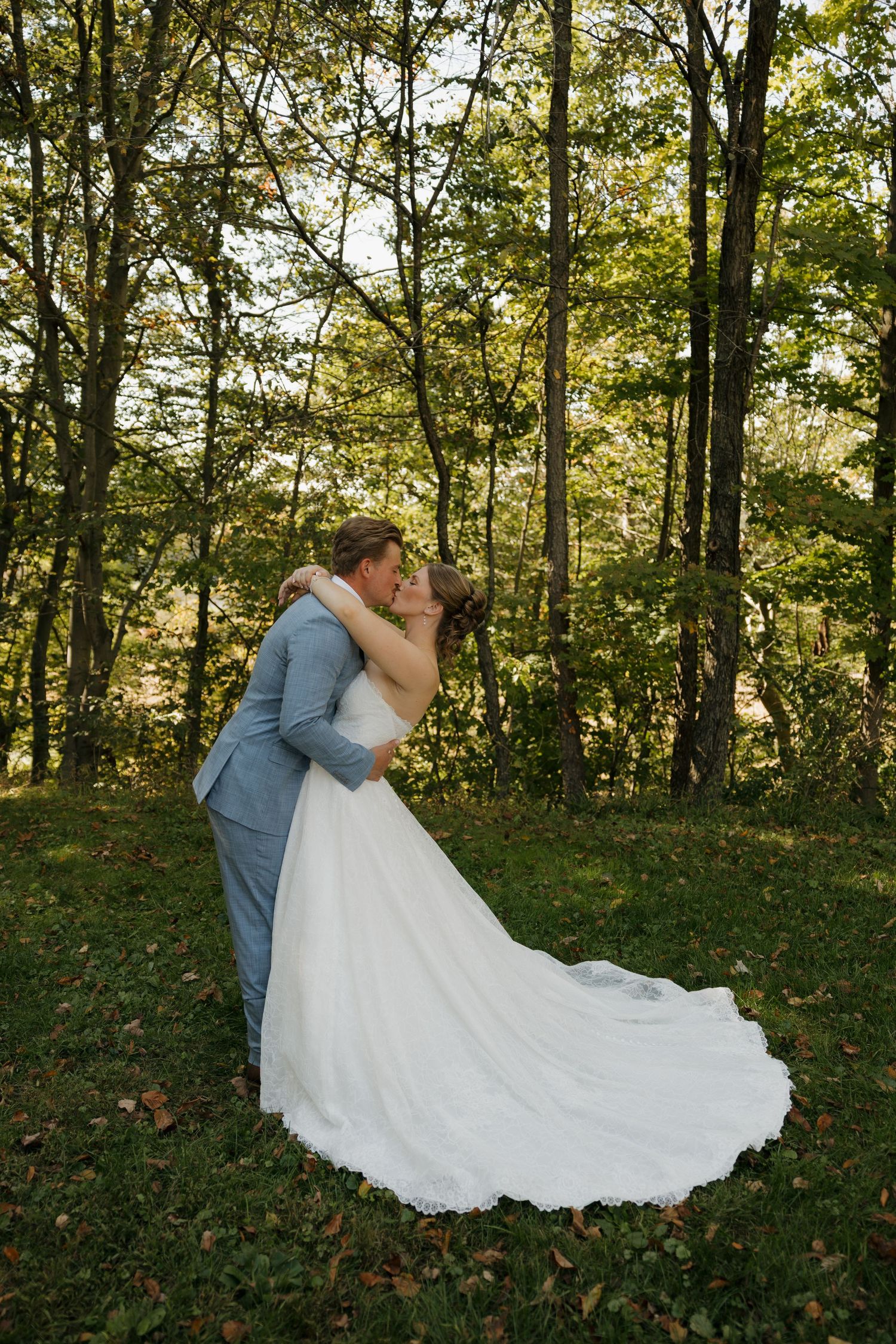 Bride in a flowing white gown kisses her groom in a light blue suit on a grassy, tree-lined wedding day clearing.