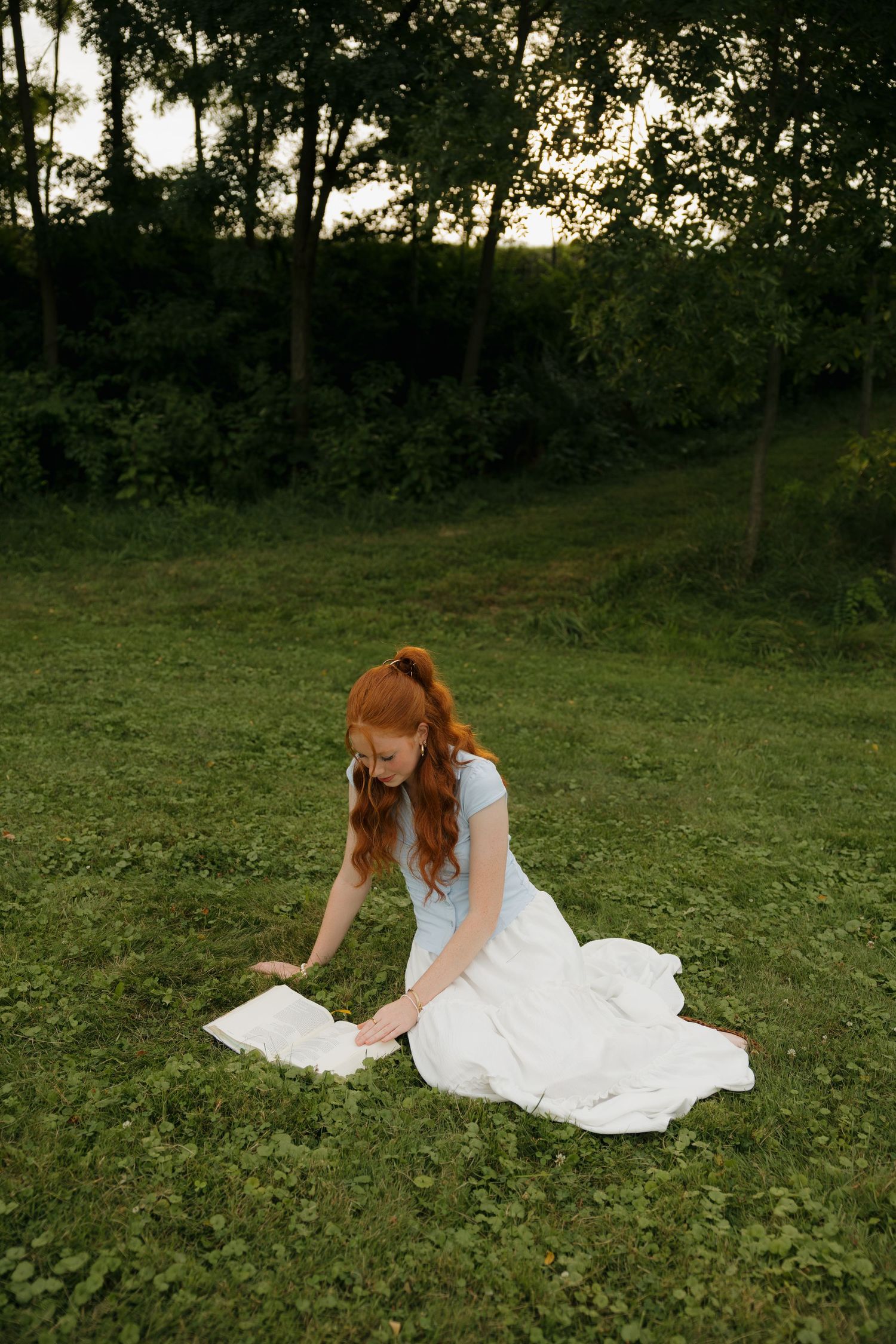 Red-haired woman in white dress reading a book on green grass at sunset with trees in background.