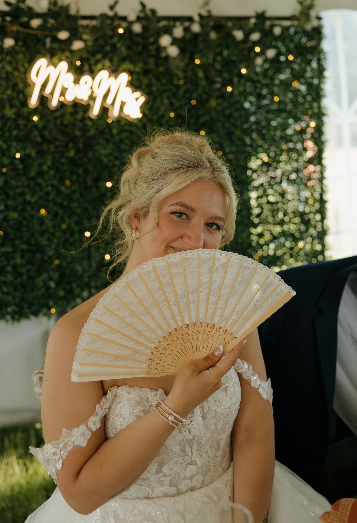 Smiling bride holding decorative white fan in front of glowing Mr & Mrs neon sign at wedding reception.