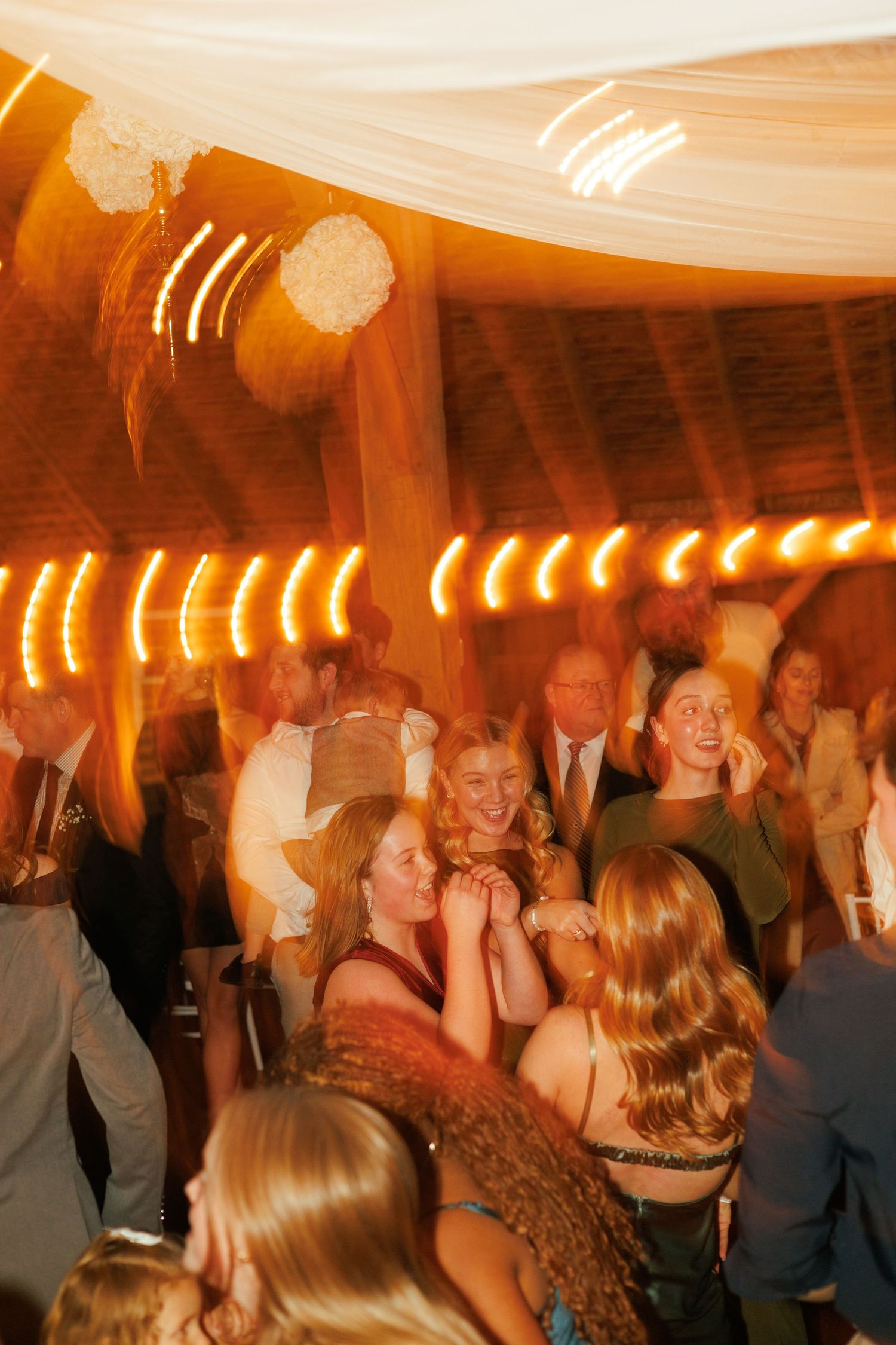 Lively dance floor scene at a wedding reception with warm lighting and white ceiling draping.
