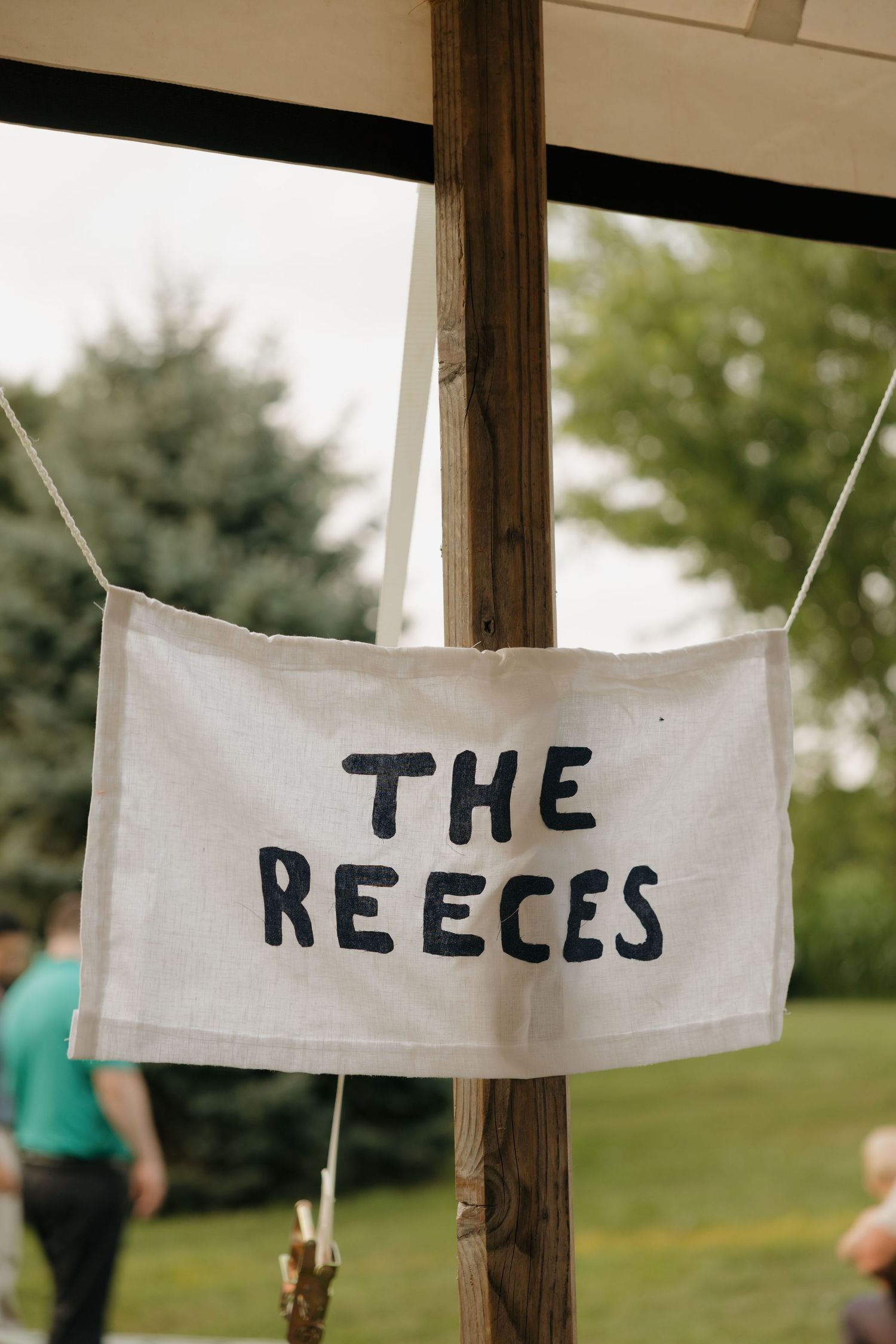 A white fabric banner reading 'The Reeces' hanging from wooden posts at an outdoor wedding reception.