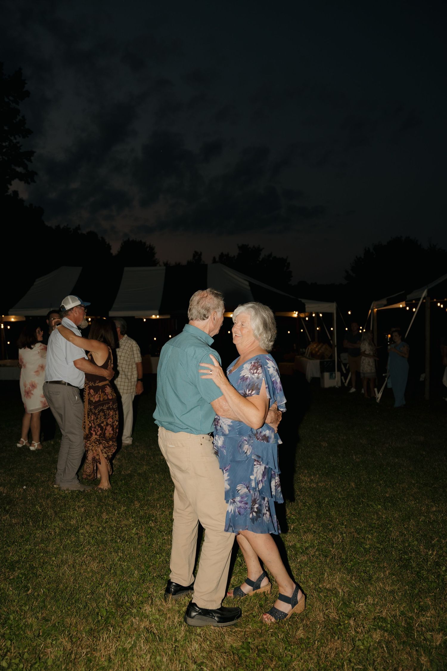 Elderly couple dances together under the night sky at an outdoor wedding reception with string lights and tent.