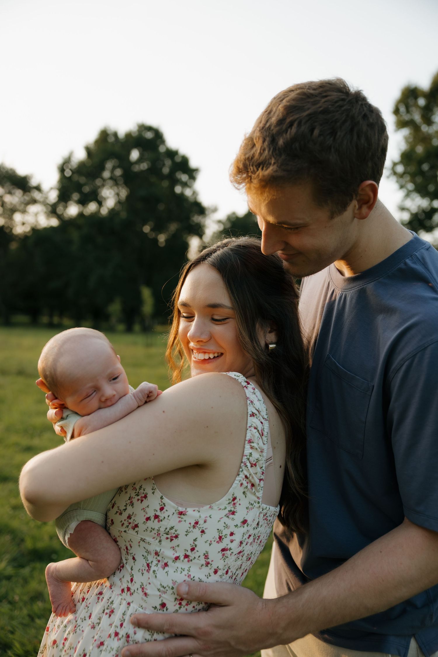 Young couple smiling outdoors at golden hour, lovingly holding their newborn baby in a green park setting.