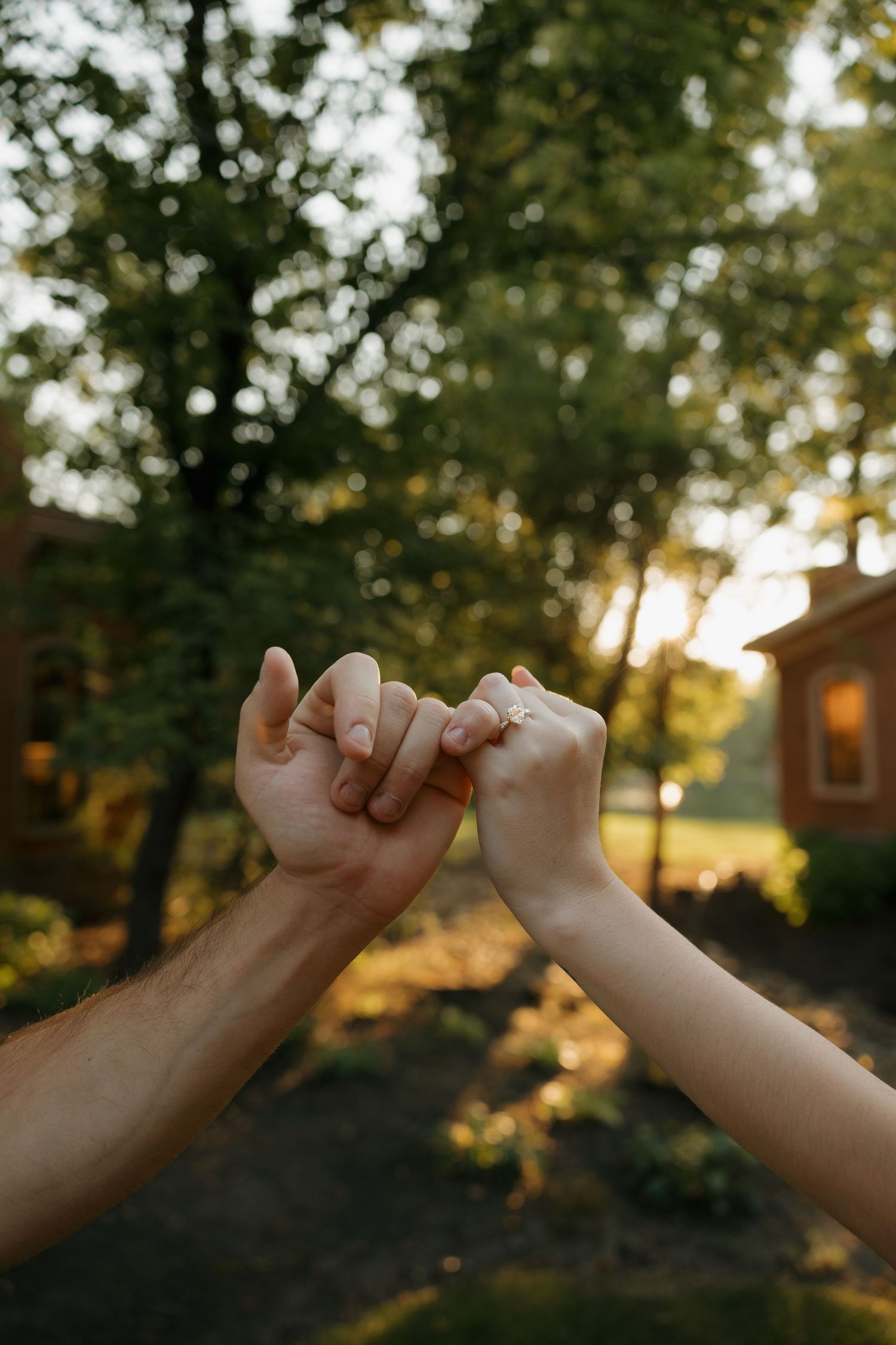 Two people share a tender pinky promise gesture outdoors at golden hour with lush green trees in the background.