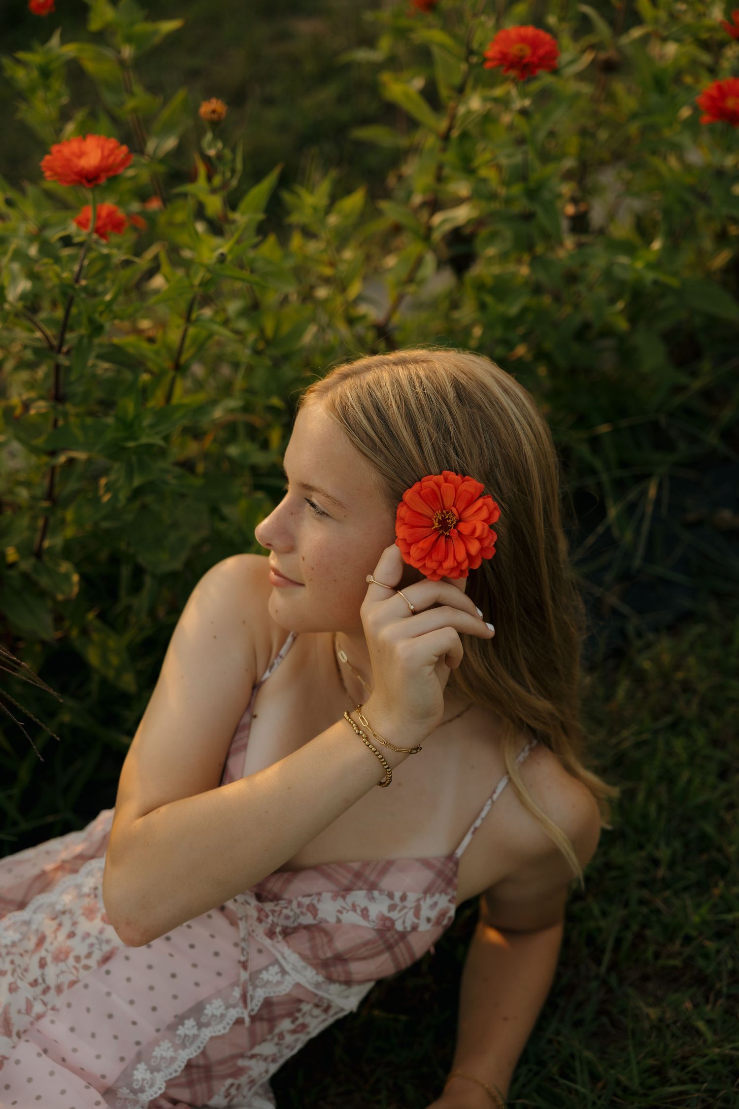 Young woman in floral dress holding red zinnia flower near her face while sitting in a garden at golden hour.