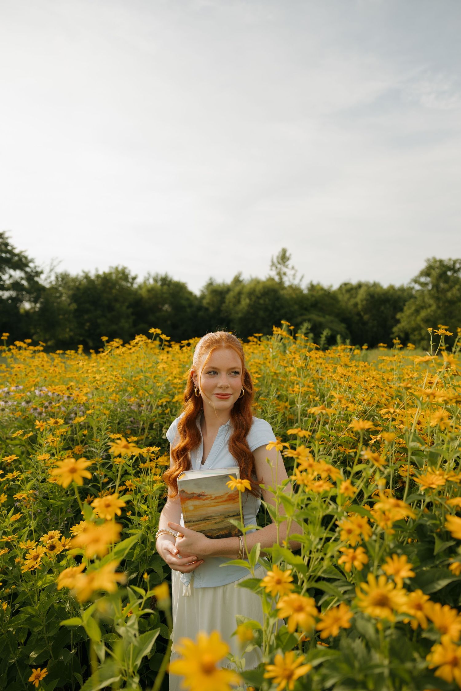 Red-haired woman in white dress holding a book in a vibrant yellow wildflower field with green trees behind.