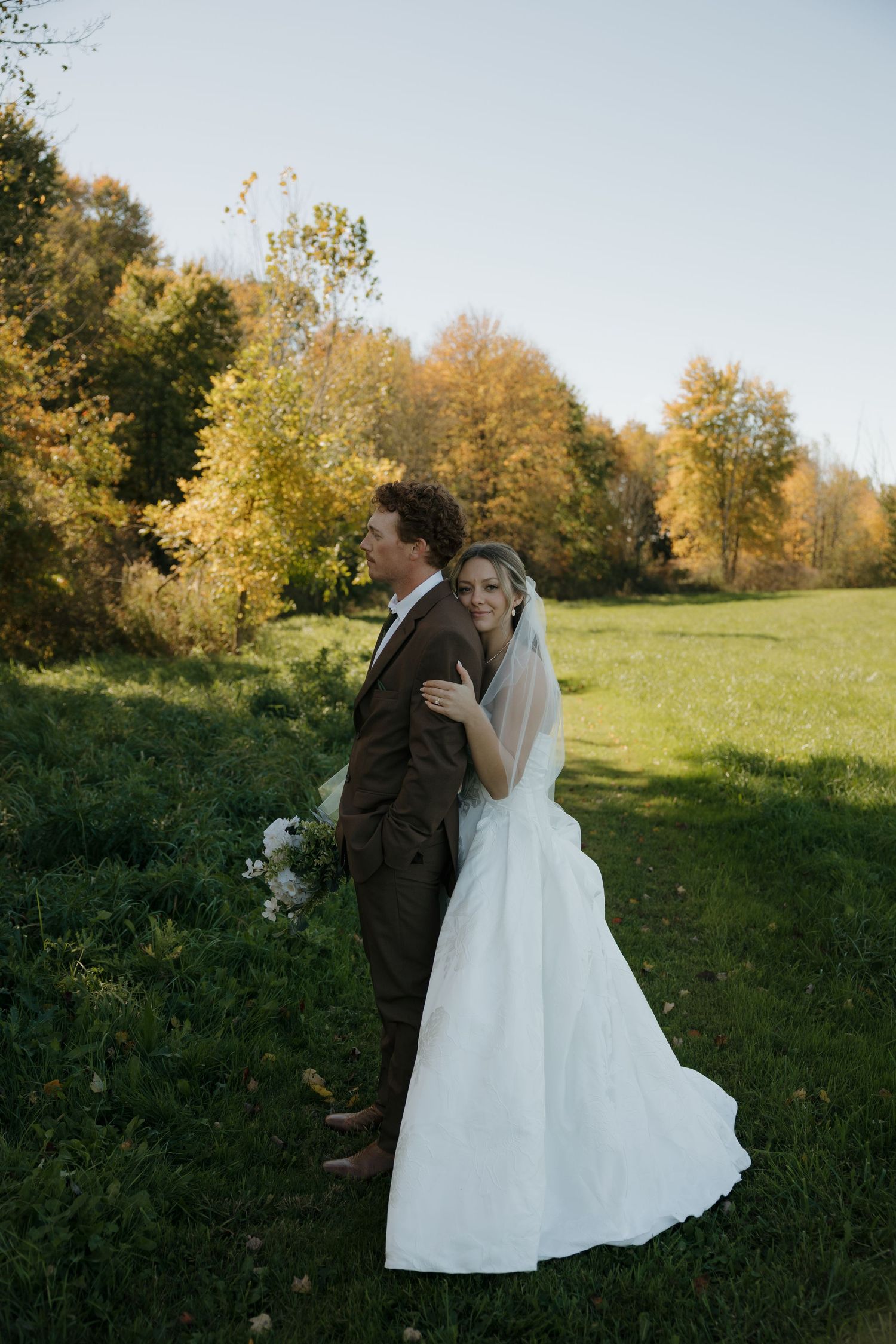 Bride in a white gown and veil embraces her groom in a brown tweed suit in a sunny meadow framed by golden autumn trees.