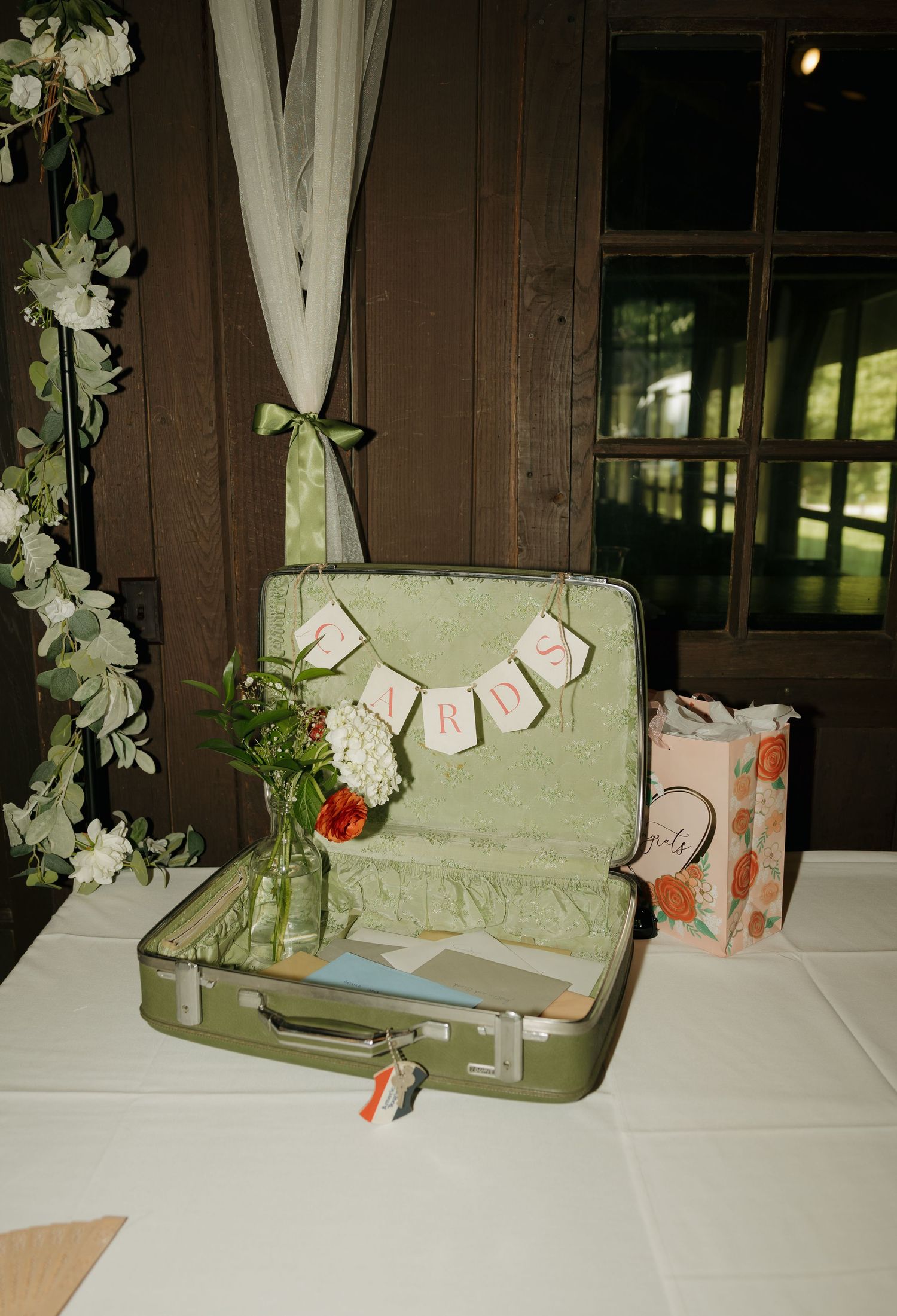 Vintage green suitcase wedding card holder with bunting banner on white table, decorated with floral arrangements at reception.
