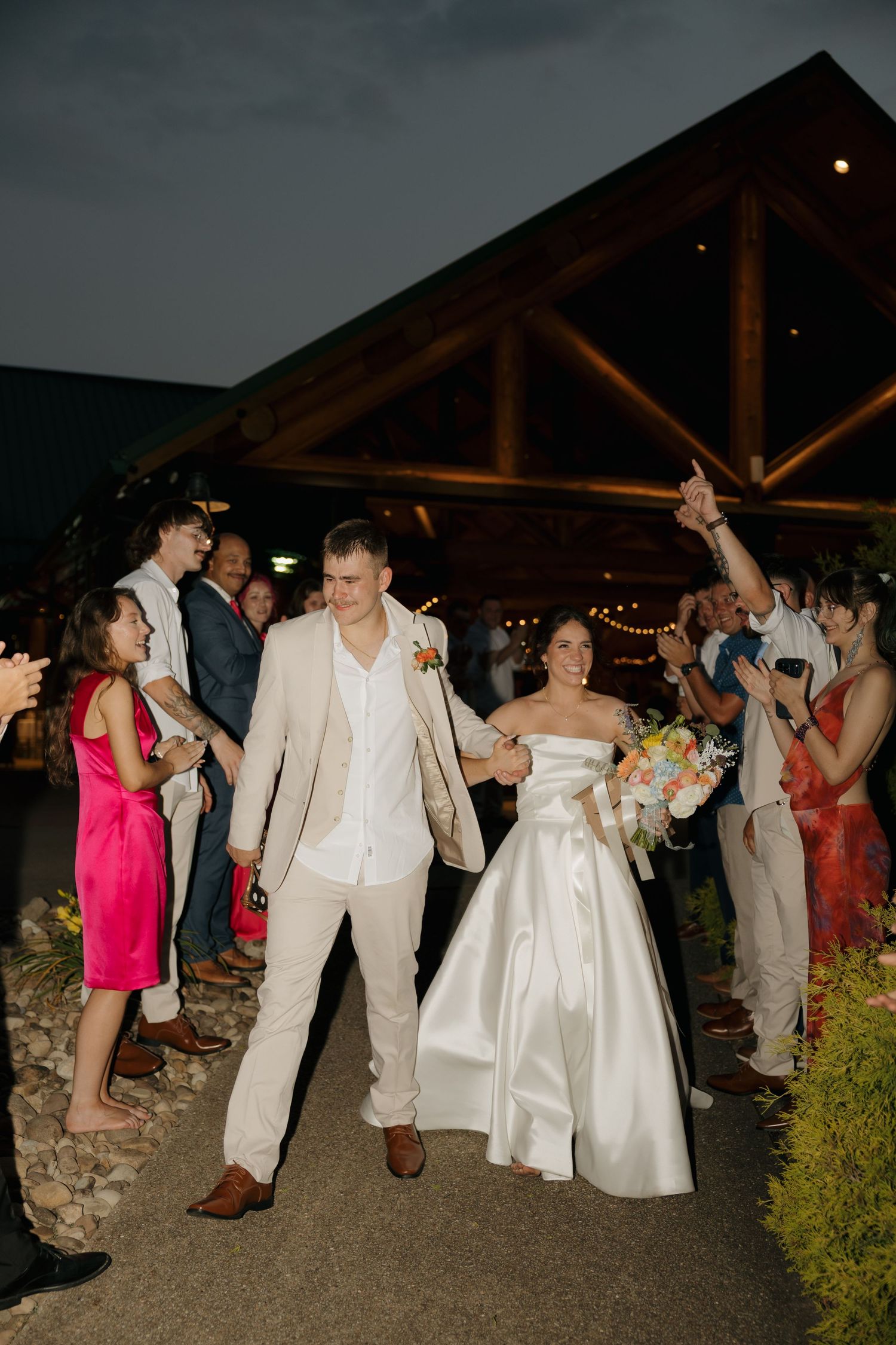 Bride and groom exit wedding reception at night, guests cheering, bride holding colorful bouquet, groom in tan suit.