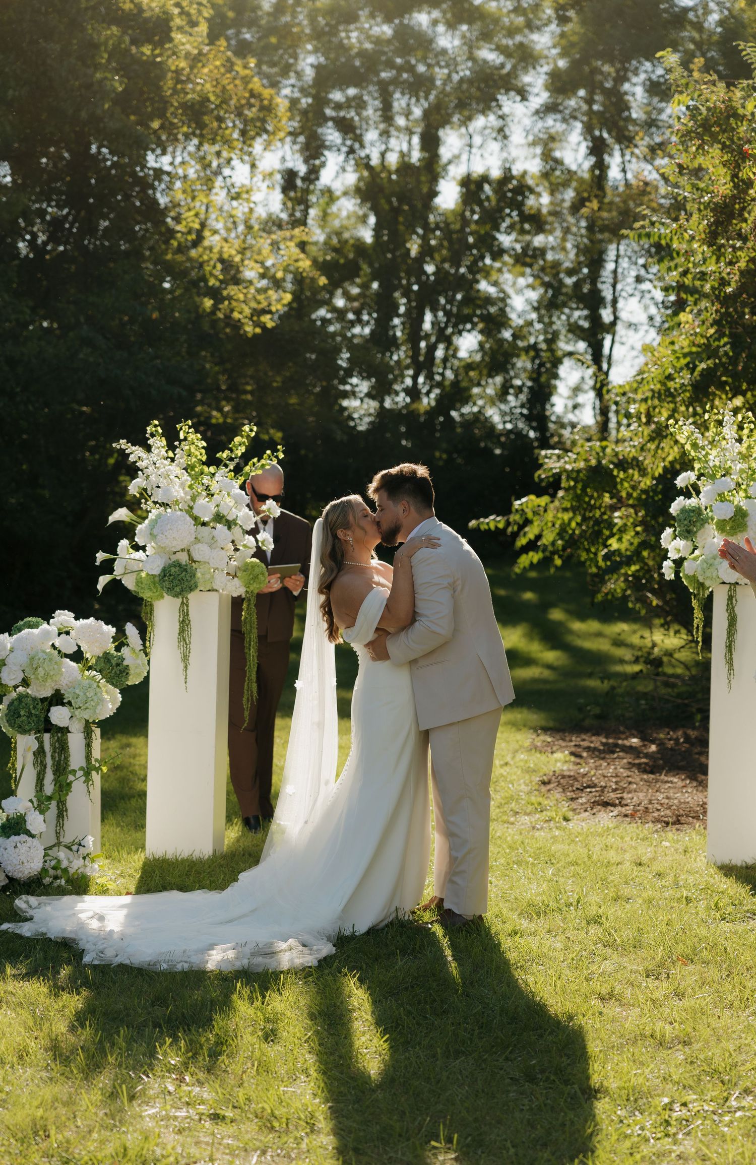 Bride and groom share first kiss at outdoor wedding ceremony surrounded by white floral arrangements on pedestals.