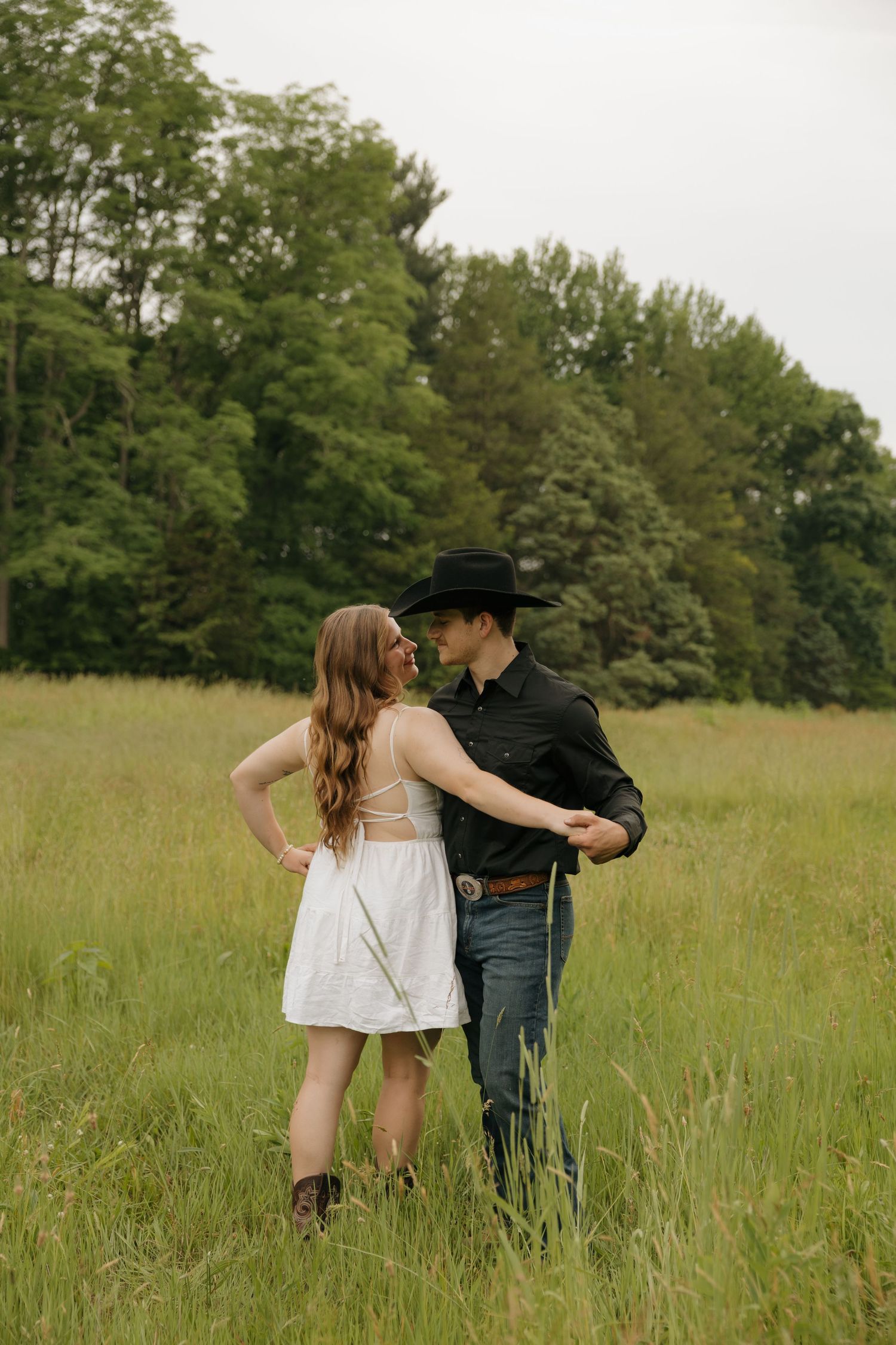 Couple dancing in a green meadow, woman in white dress and cowboy boots, man in black shirt and cowboy hat.