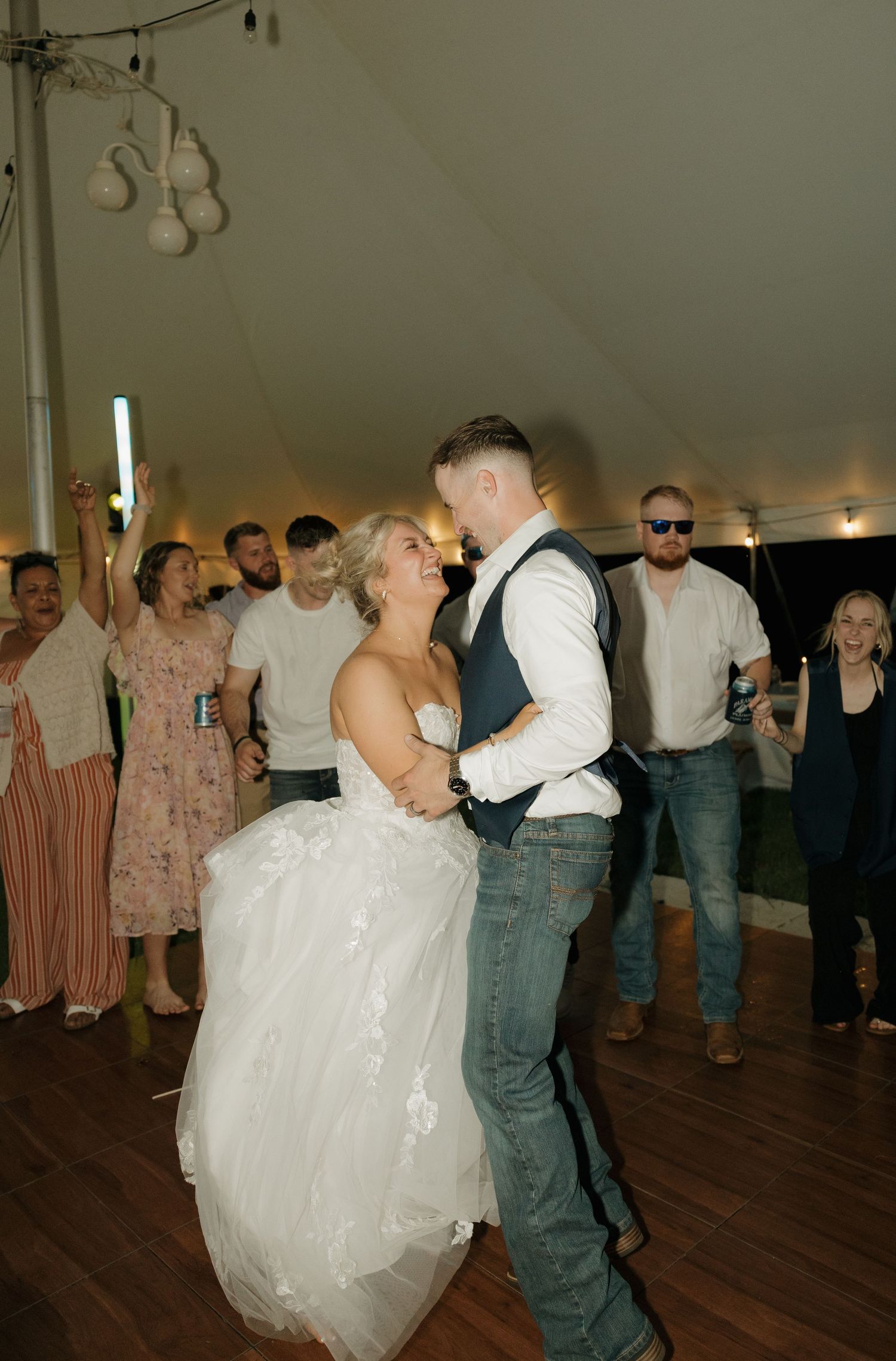 Bride and groom share first dance at wedding reception in white tent, surrounded by cheering guests.