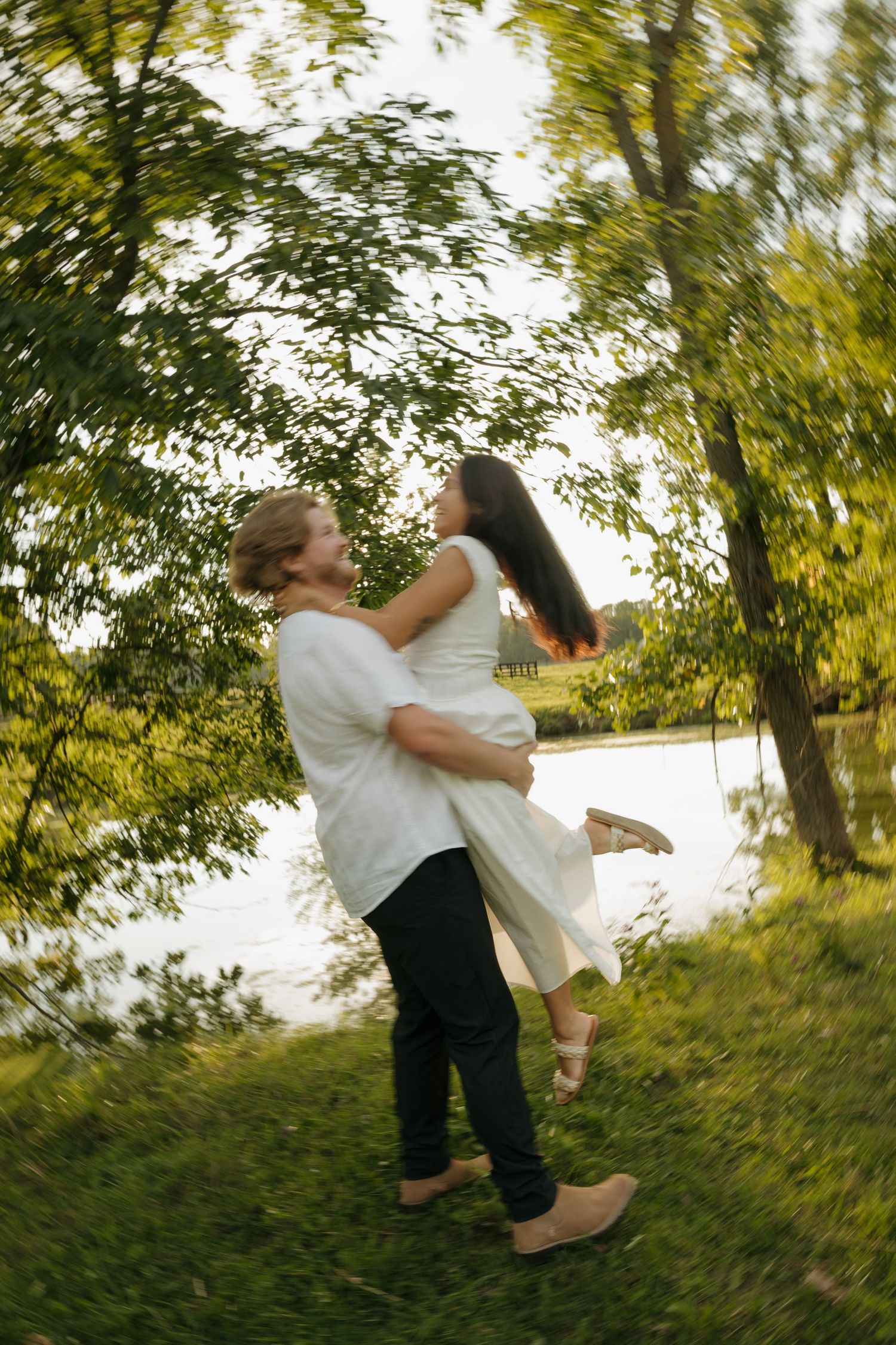 A joyful couple embraces and spins together in a lush green park near a lake, captured with a dreamy motion blur effect.