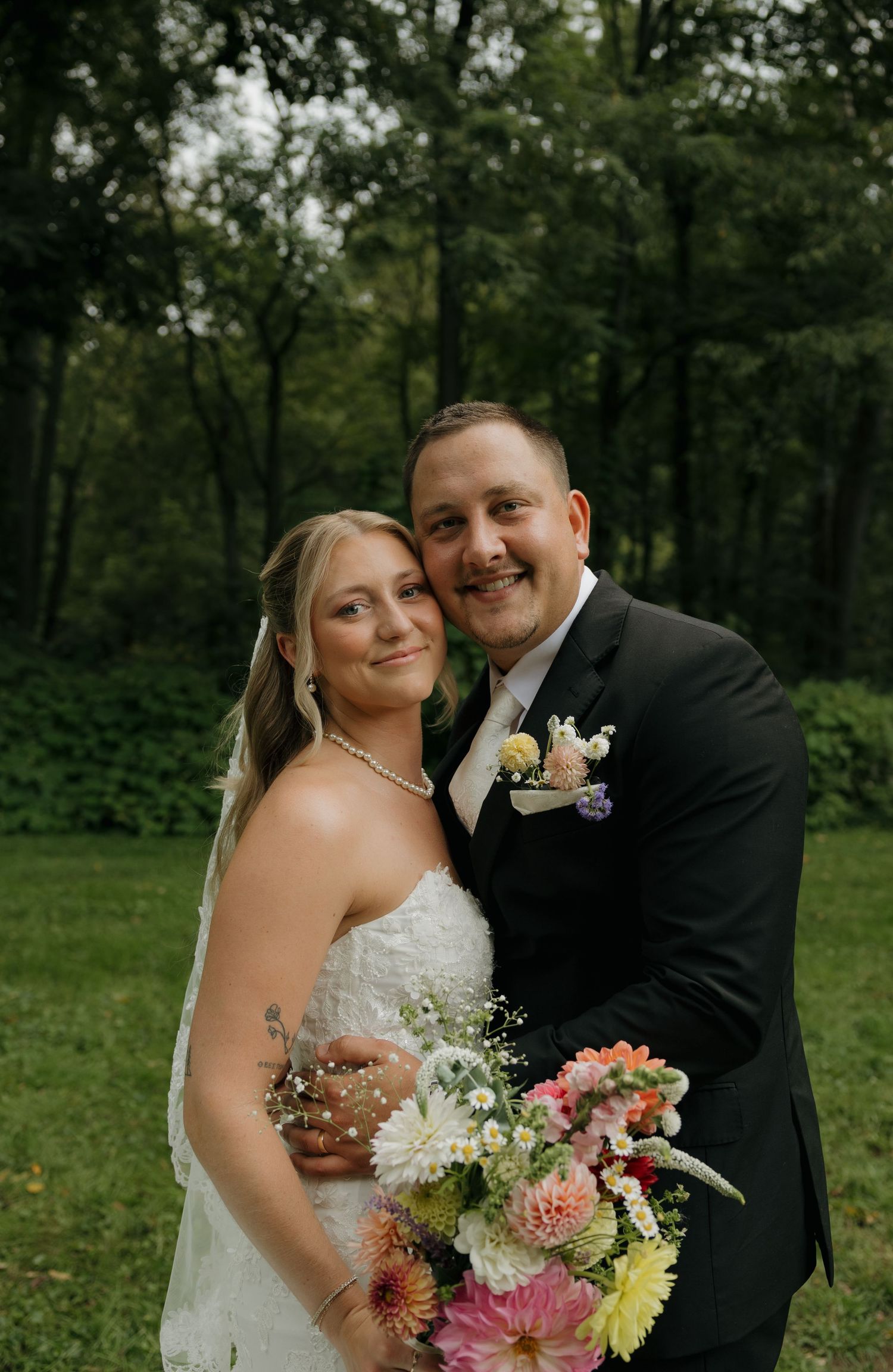 A smiling bride and groom pose together outdoors in a lush green setting, she holding a colorful bouquet on their wedding day.