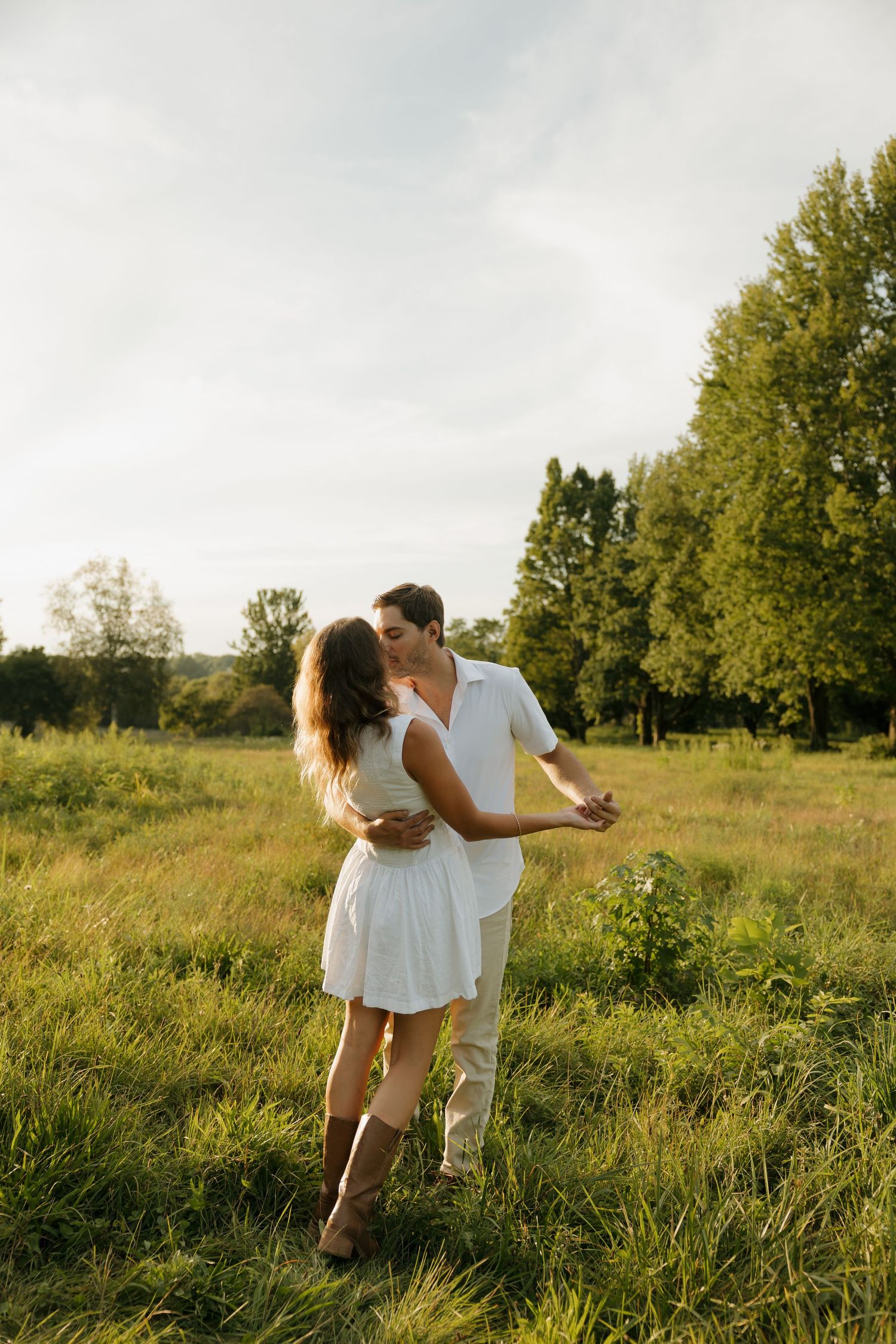 Romantic couple in white outfits dancing and kissing in a sunlit green meadow with trees in the background.