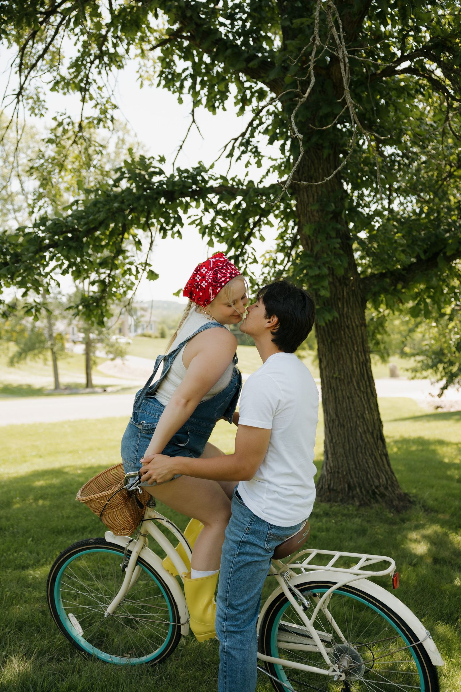 Couple sharing a romantic kiss in a park, with woman sitting on a vintage white bicycle under a large green tree.
