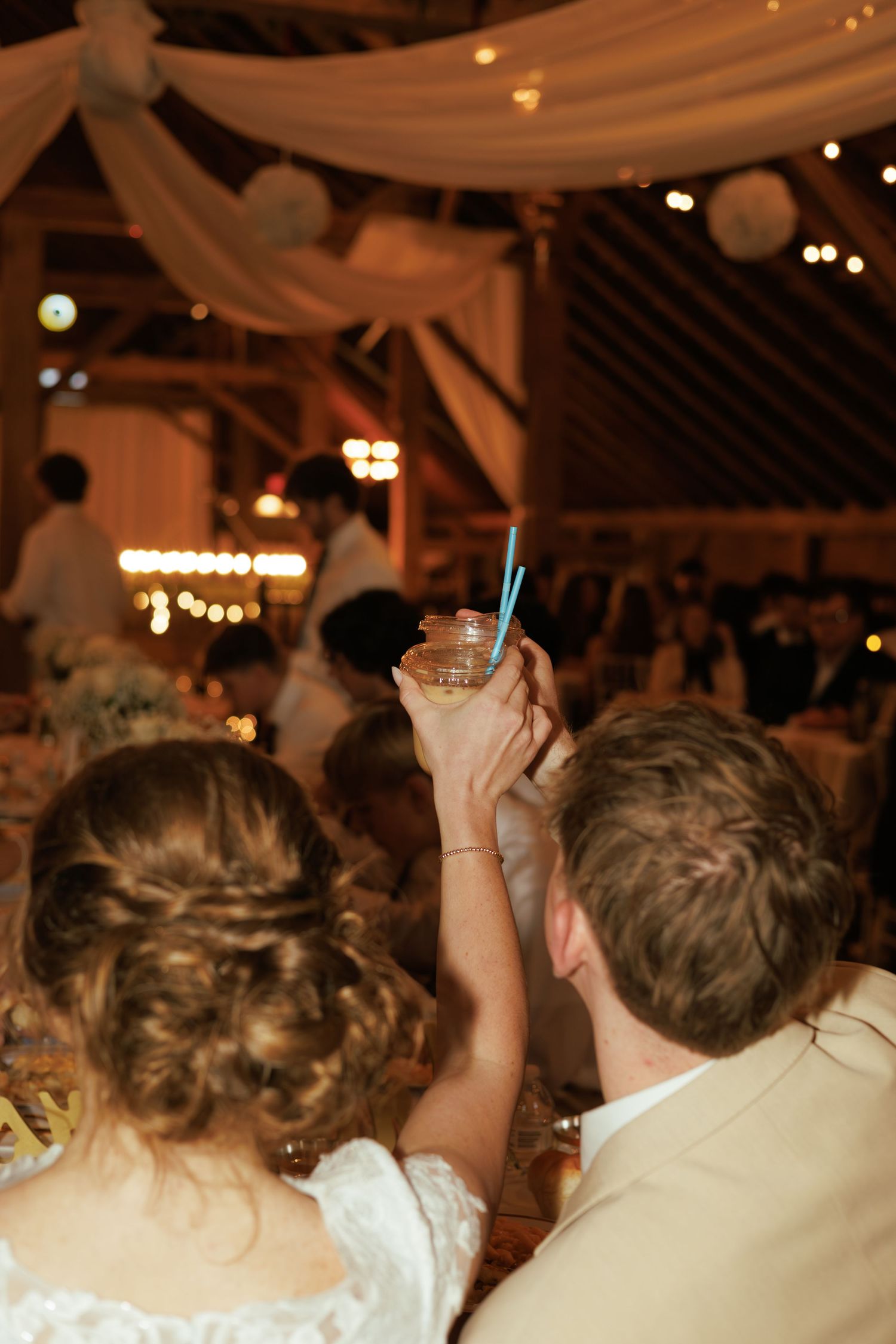 Bride and groom raising a drink in a rustic barn wedding reception with warm string lights and draped fabric.