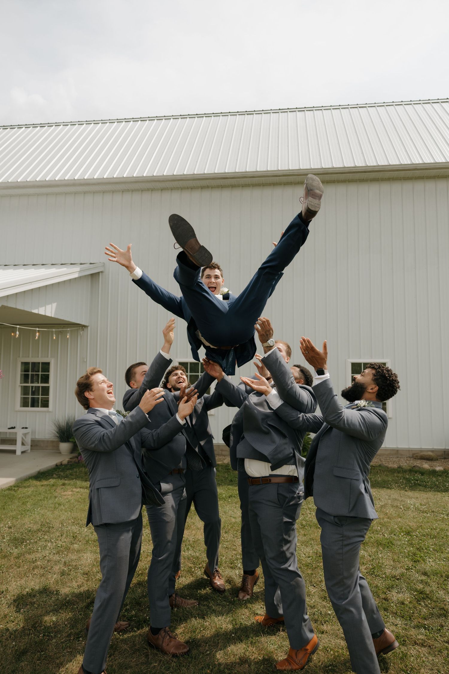 Groomsmen toss the groom into the air in a celebratory moment outside a white barn venue.