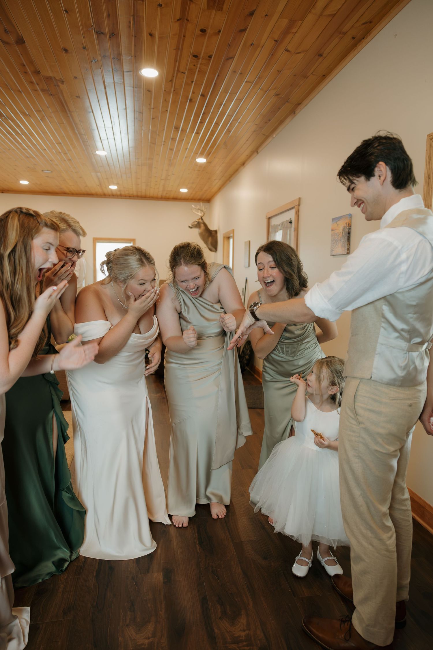 Bridesmaids in sage green dresses laugh with a groom and flower girl during wedding preparations indoors.