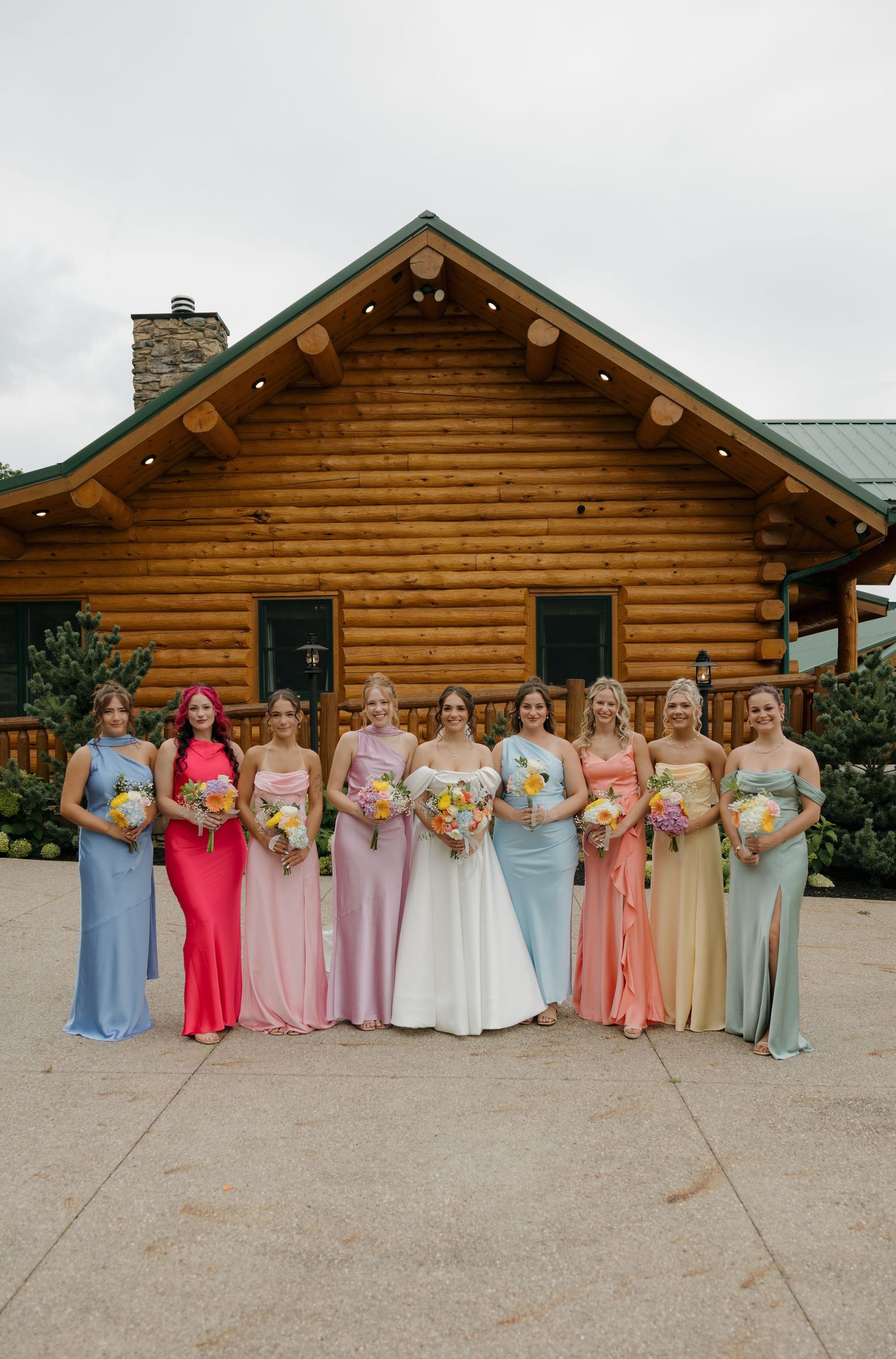 Bride and bridesmaids in rainbow-colored gowns holding bouquets outside a rustic log cabin venue.