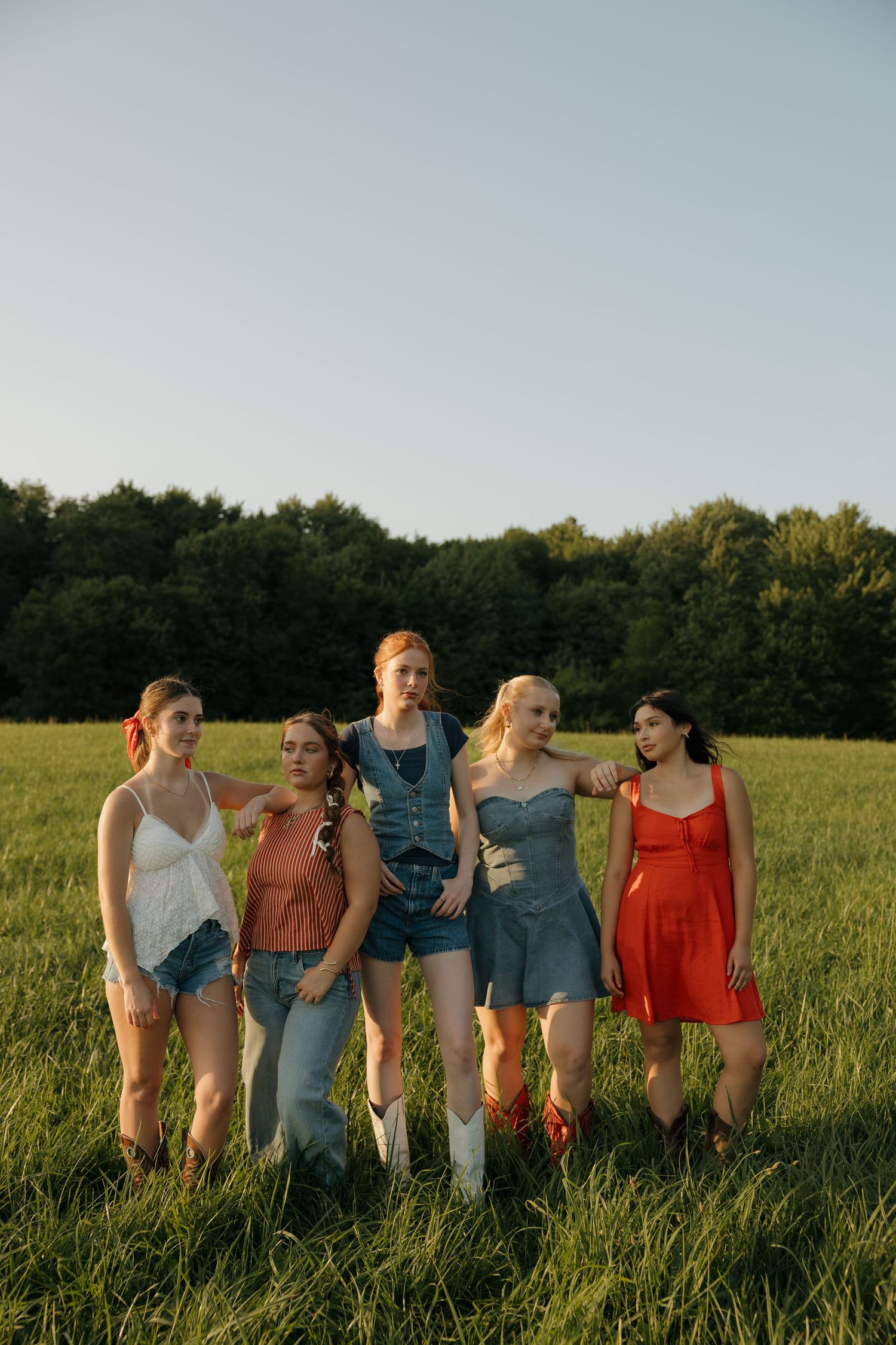 Five young women pose together in a green field at golden hour, wearing summer outfits including denim and red dresses.