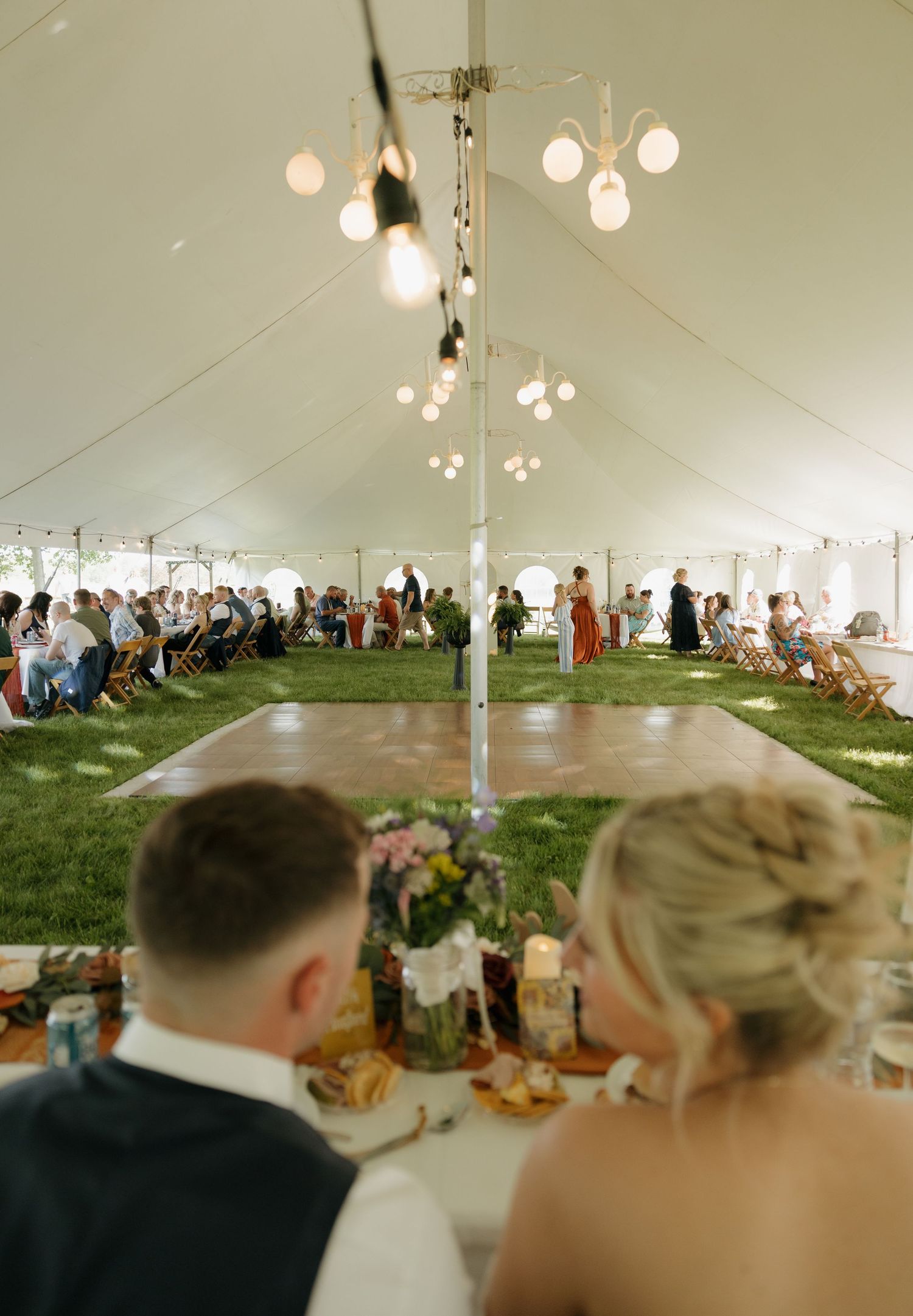 Aerial view of elegant outdoor tent wedding reception with dance floor, string lights, and guests seated on wooden chairs.