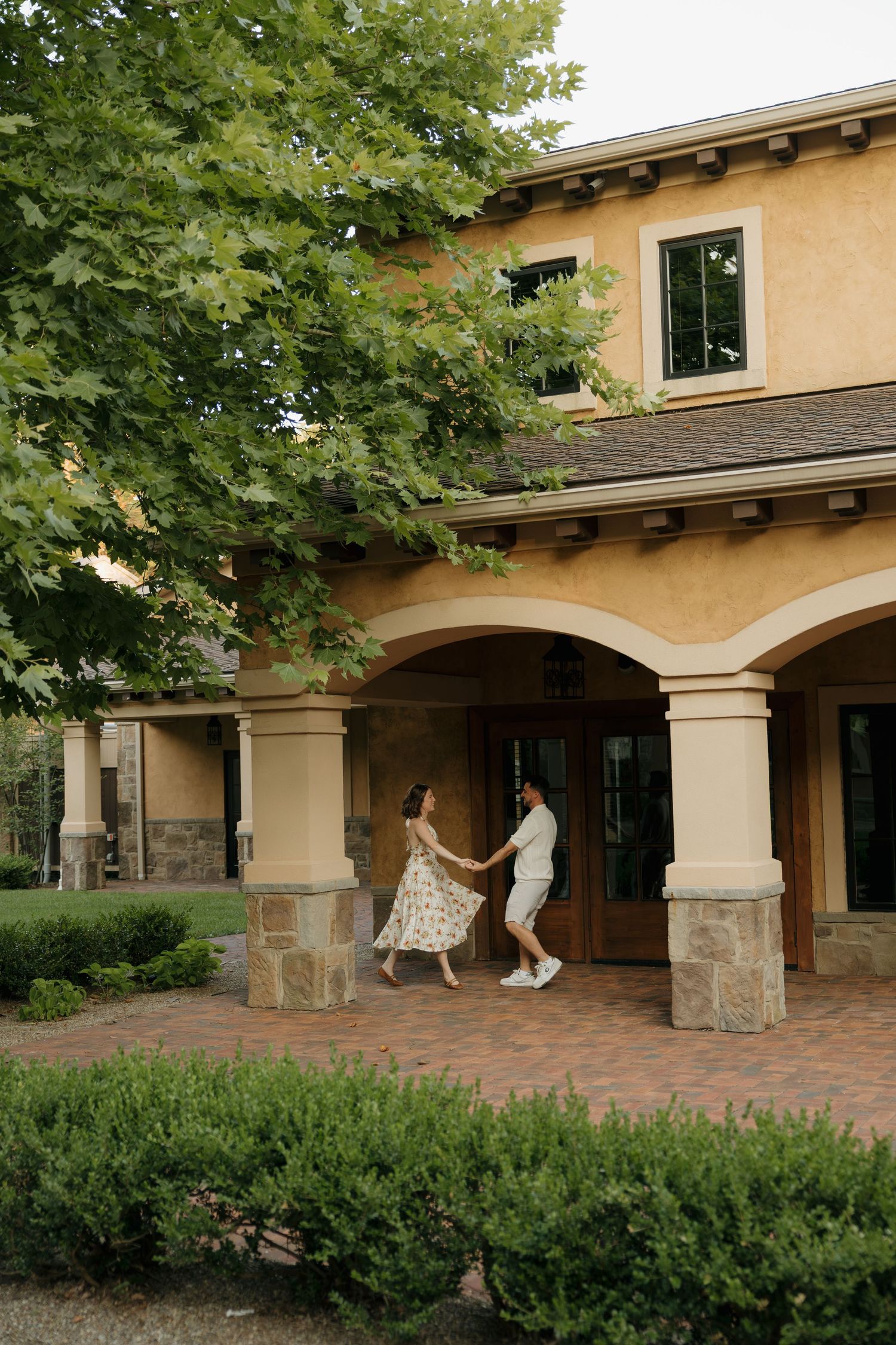 A couple dances joyfully on a brick patio outside a elegant Mediterranean-style villa surrounded by lush greenery.