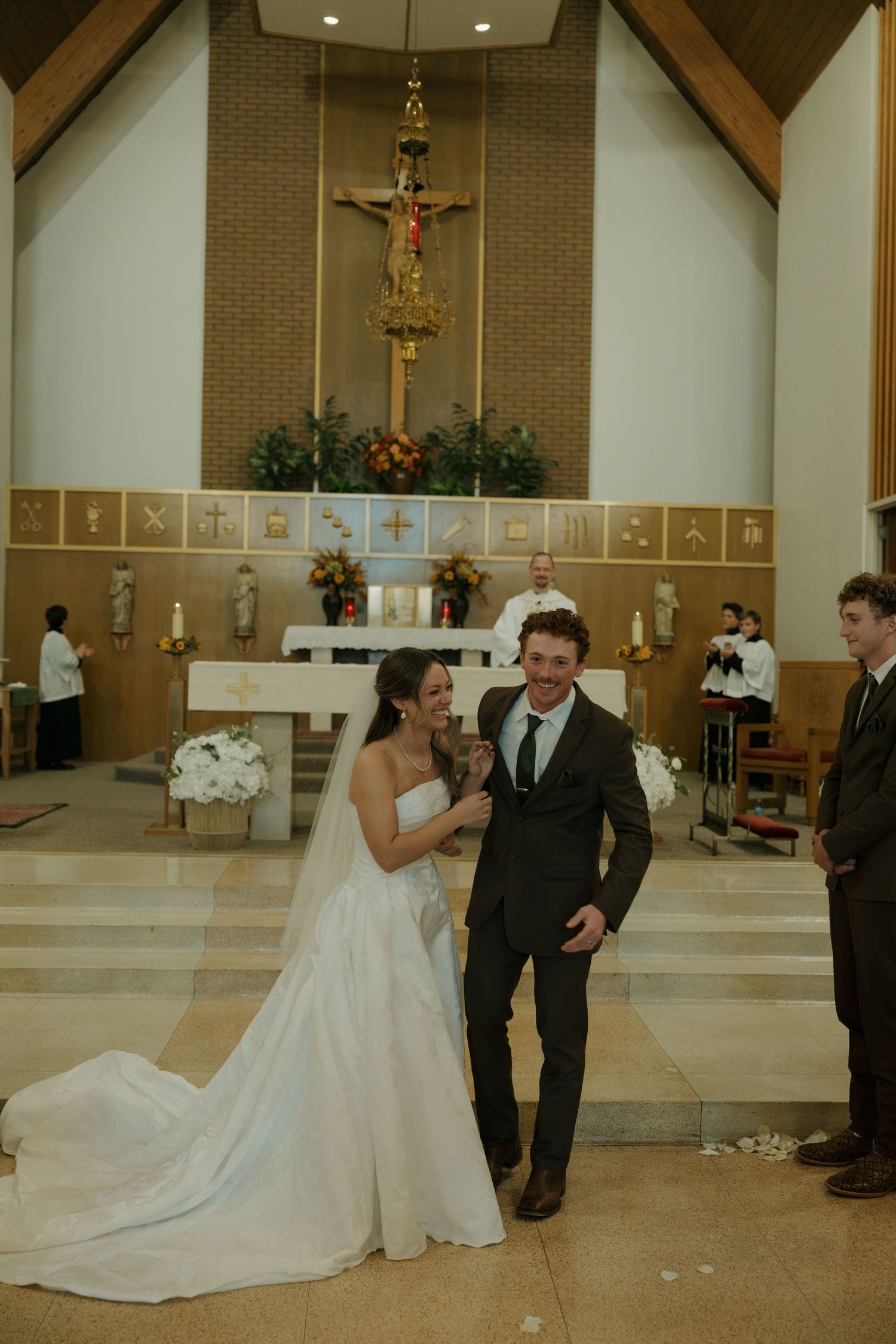 Bride in a long-veiled gown laughs with her groom at the church altar during their wedding ceremony, with a crucifix behind them.