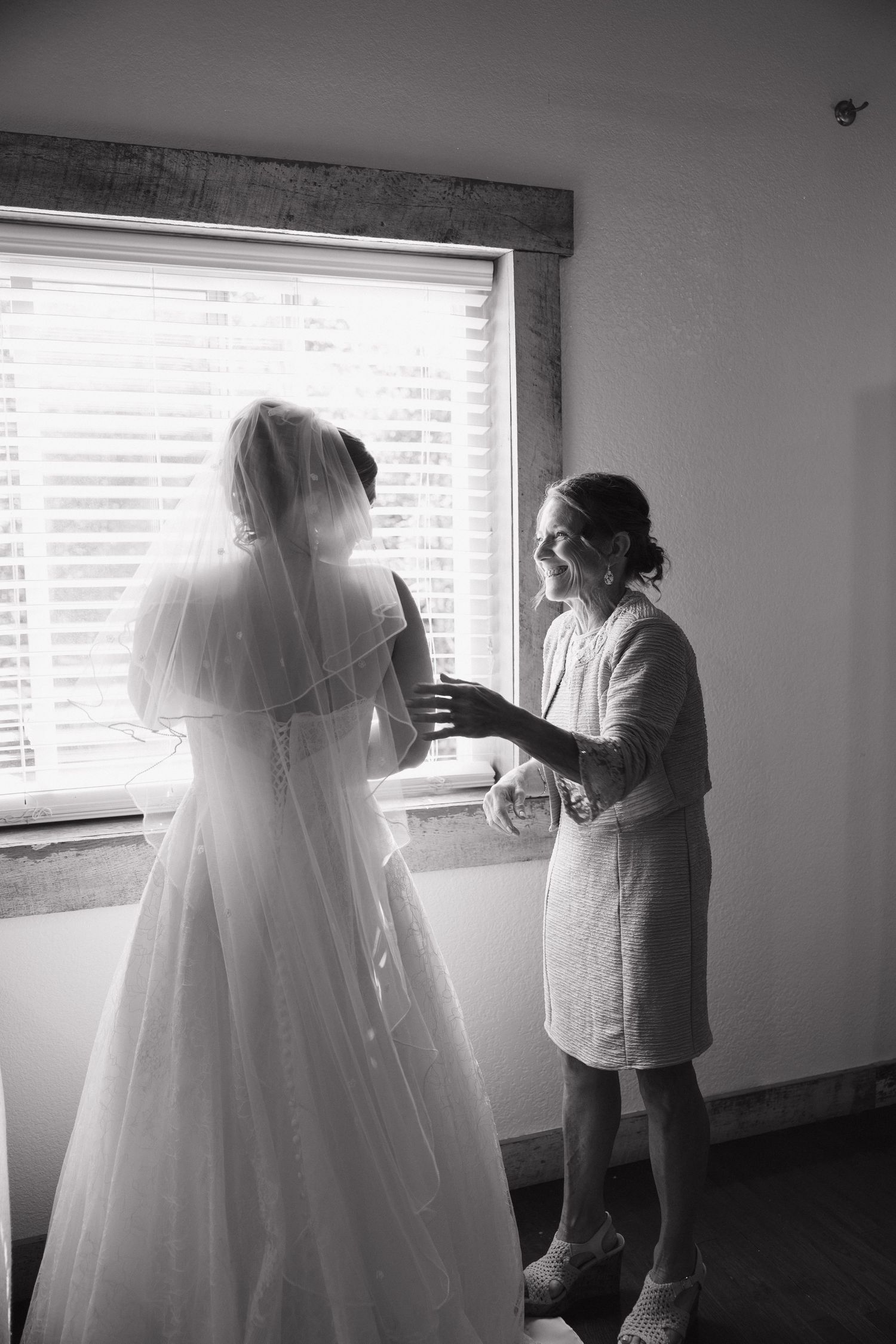 Black and white photo of a bride in a long veil and gown by a window as a smiling woman in a tweed jacket helps her get ready.