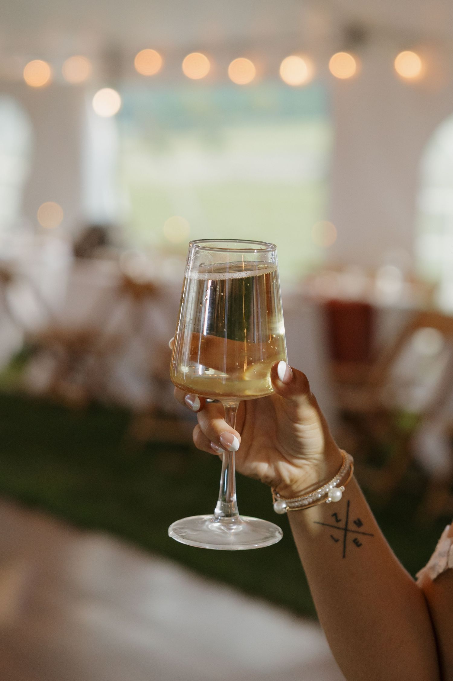 A hand holding a champagne flute at an elegant wedding reception tent with string lights in the background.