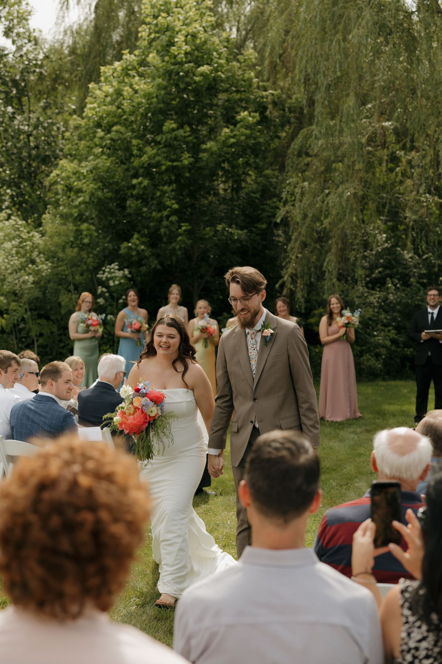 Bride and groom walk down outdoor aisle holding hands, bride carries colorful bouquet, guests celebrate in lush green garden setting.