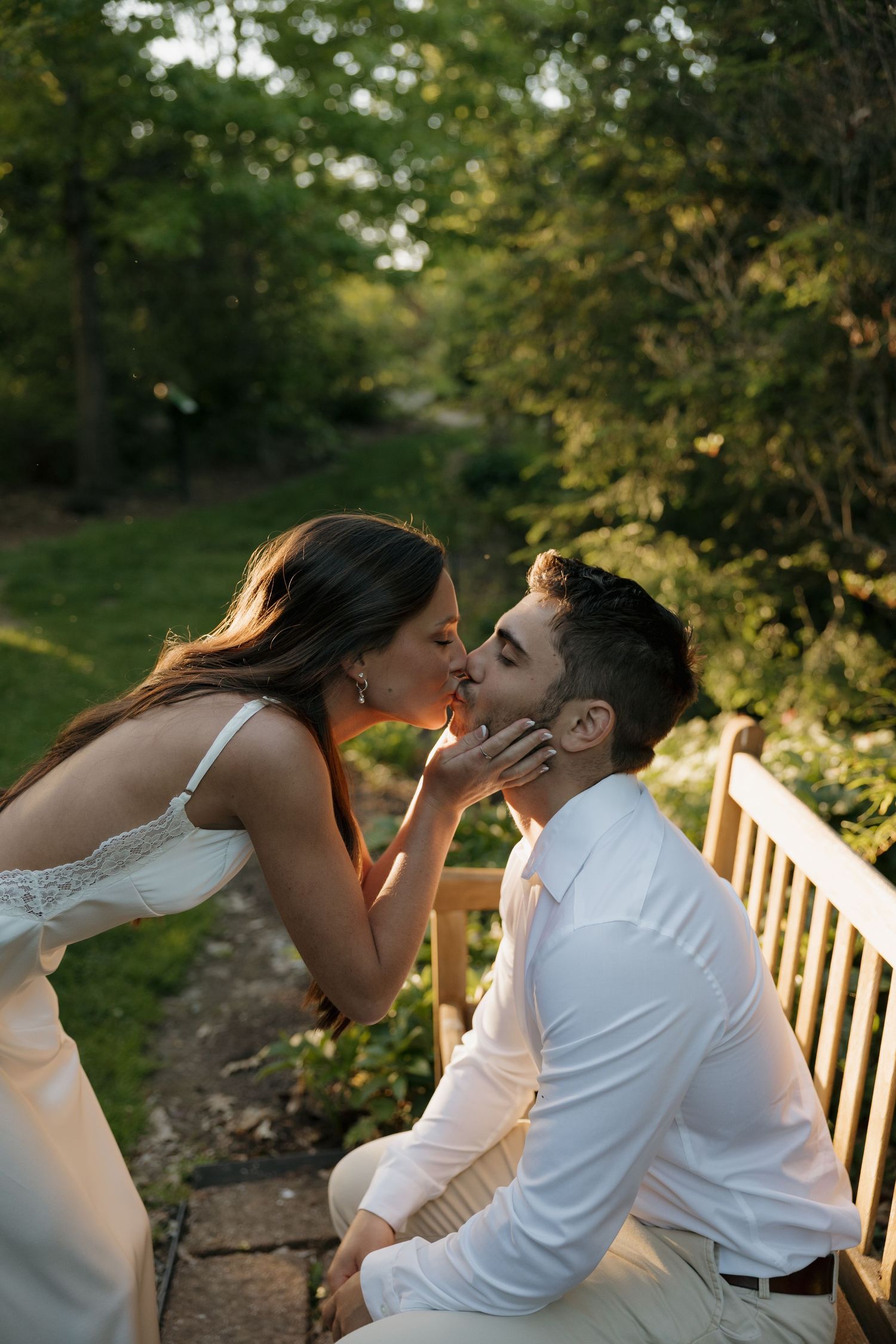 A couple shares a romantic kiss on a wooden bench surrounded by lush green trees in a garden setting.