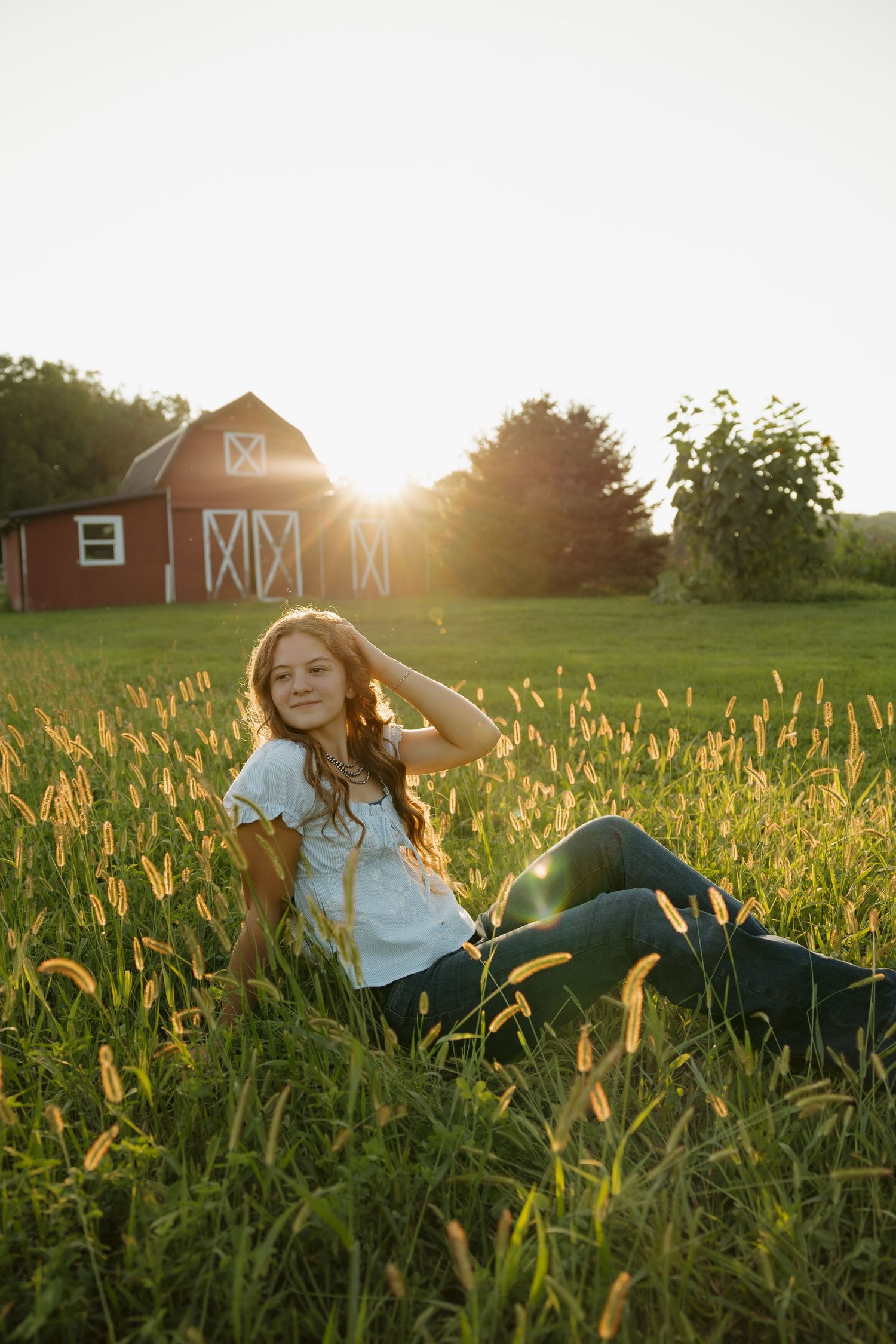 Young woman sitting in golden grass at sunset with a red barn glowing in the background on a farm.