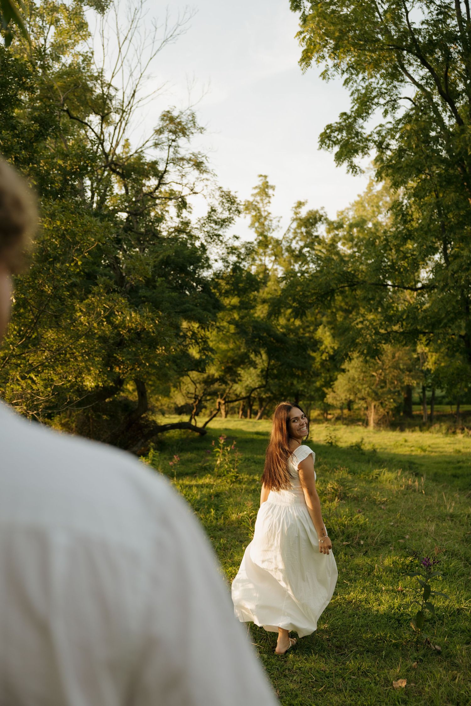 Woman in white dress walking barefoot on green grass in a sunlit garden, looking back over her shoulder.