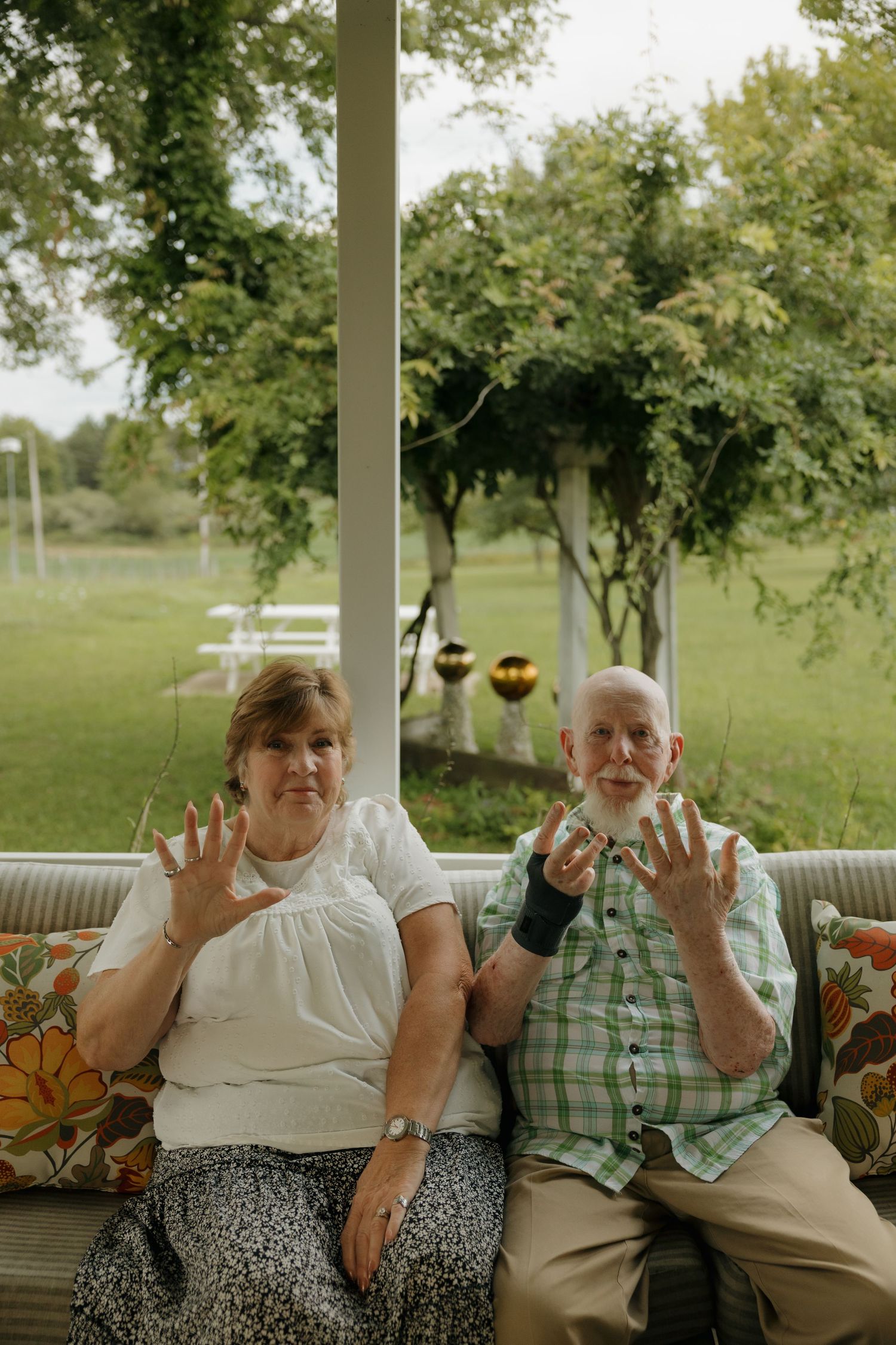 Elderly couple sitting on a sofa, raising their hands toward the camera, with a lush green garden visible behind them.