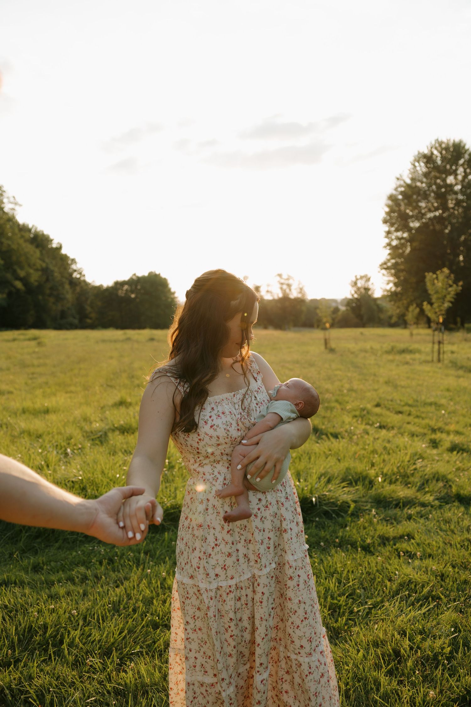 Mother in floral dress holds newborn while walking in sunlit green field at golden hour.