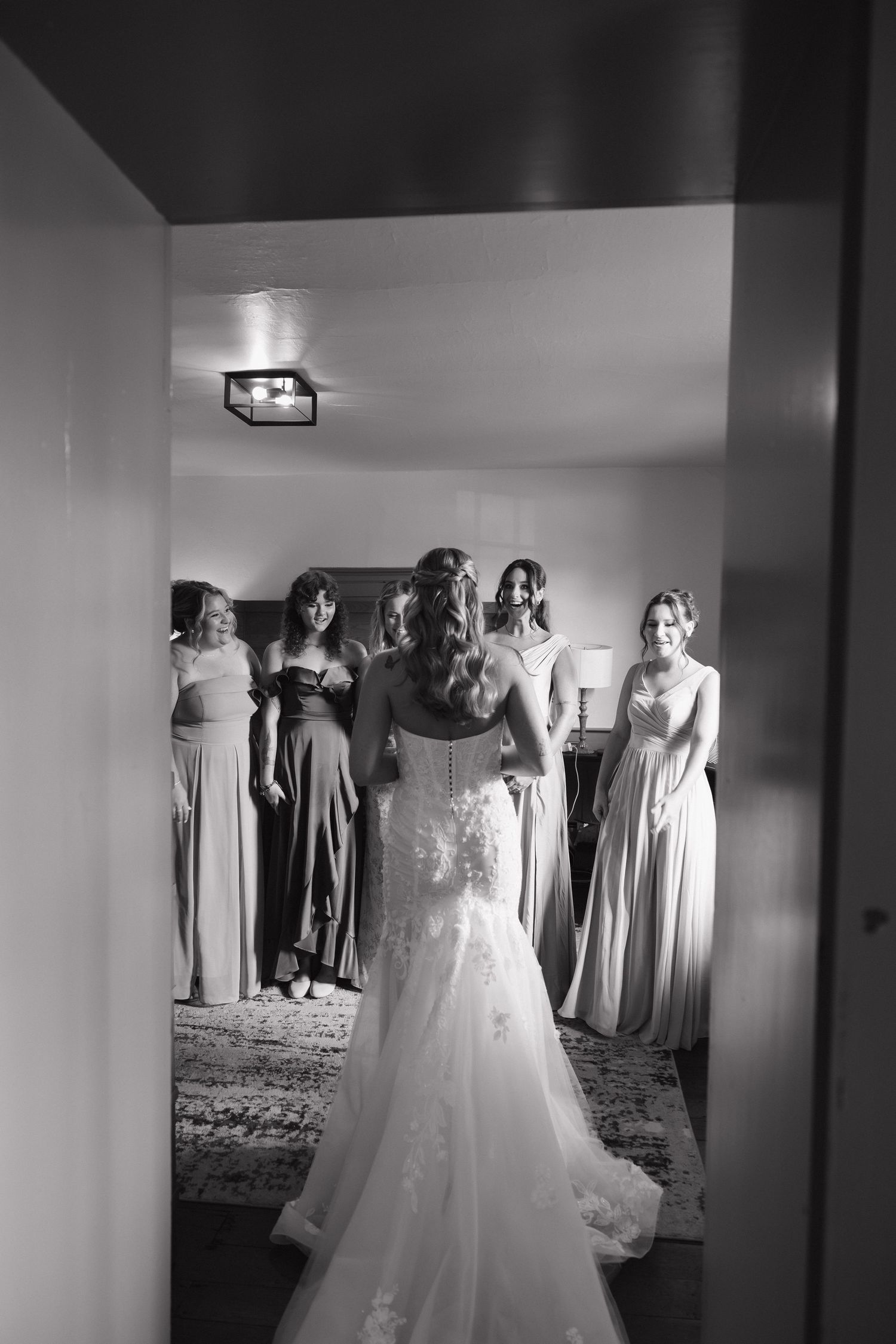 Bride in lace mermaid gown stands with bridesmaids in mismatched dresses in black and white photo.