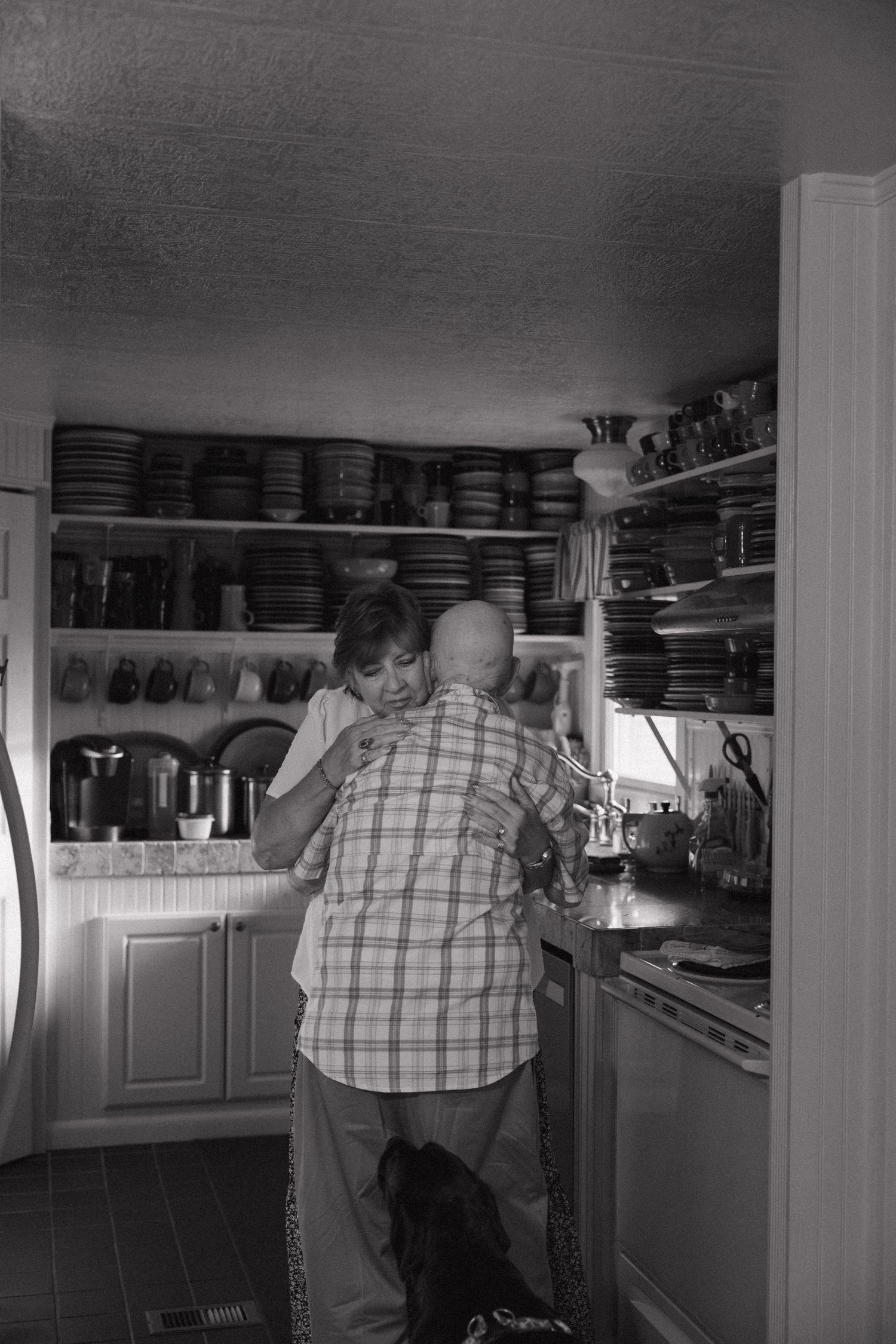 Black and white photo of a couple embracing in a cozy kitchen with shelves full of dishes and pottery.