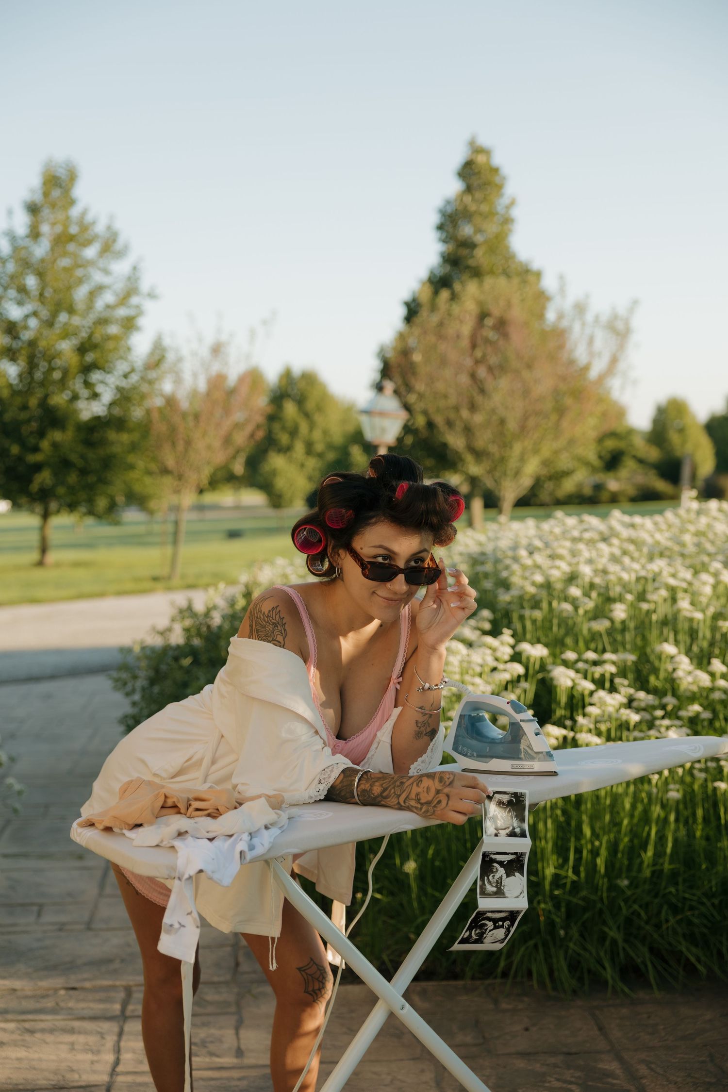 Tattooed woman with hair curlers and sunglasses leans on ironing board outdoors in a sunny park setting.