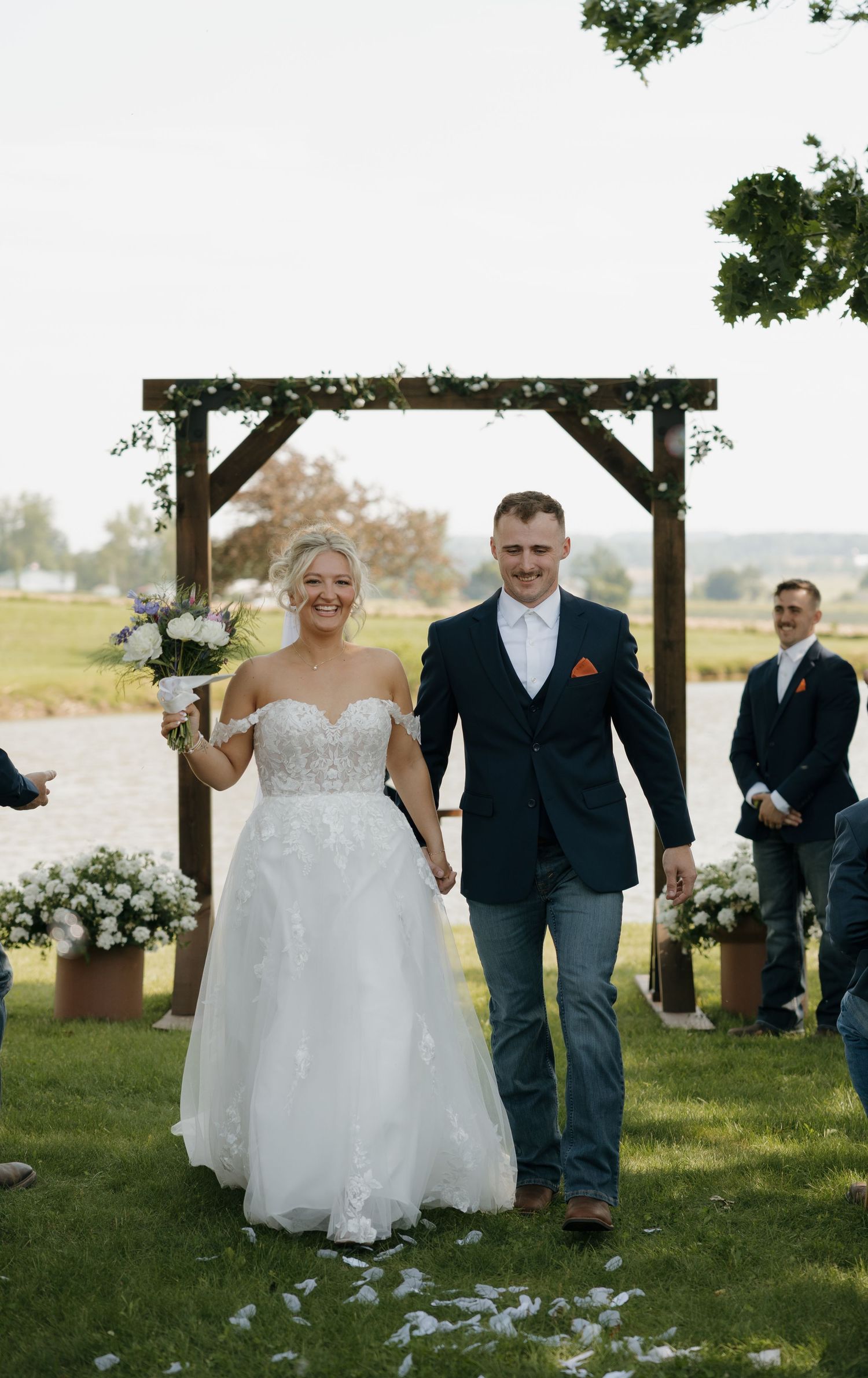 Newlyweds walking down the aisle holding hands under a rustic wooden arch by a lake.