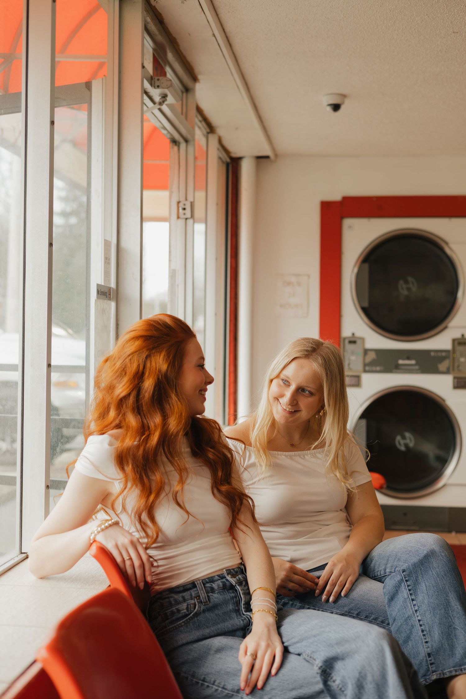 Two people in jeans and white tops lounge together near commercial washing machines in a laundromat.